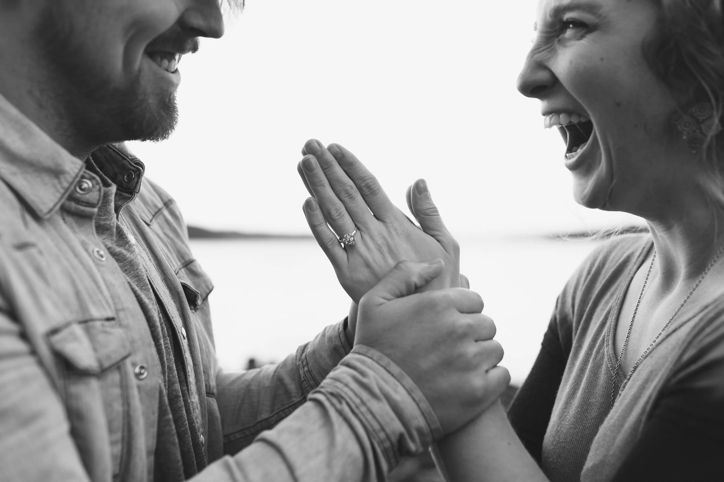 Black and white photo of a smiling couple holding hands, focusing on an engagement ring on the woman's finger.