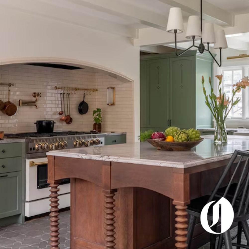 A cozy kitchen with green cabinets, a marble-topped island with wooden turned legs, a hanging light fixture, a bowl of fresh vegetables, and a vase of pink flowers.