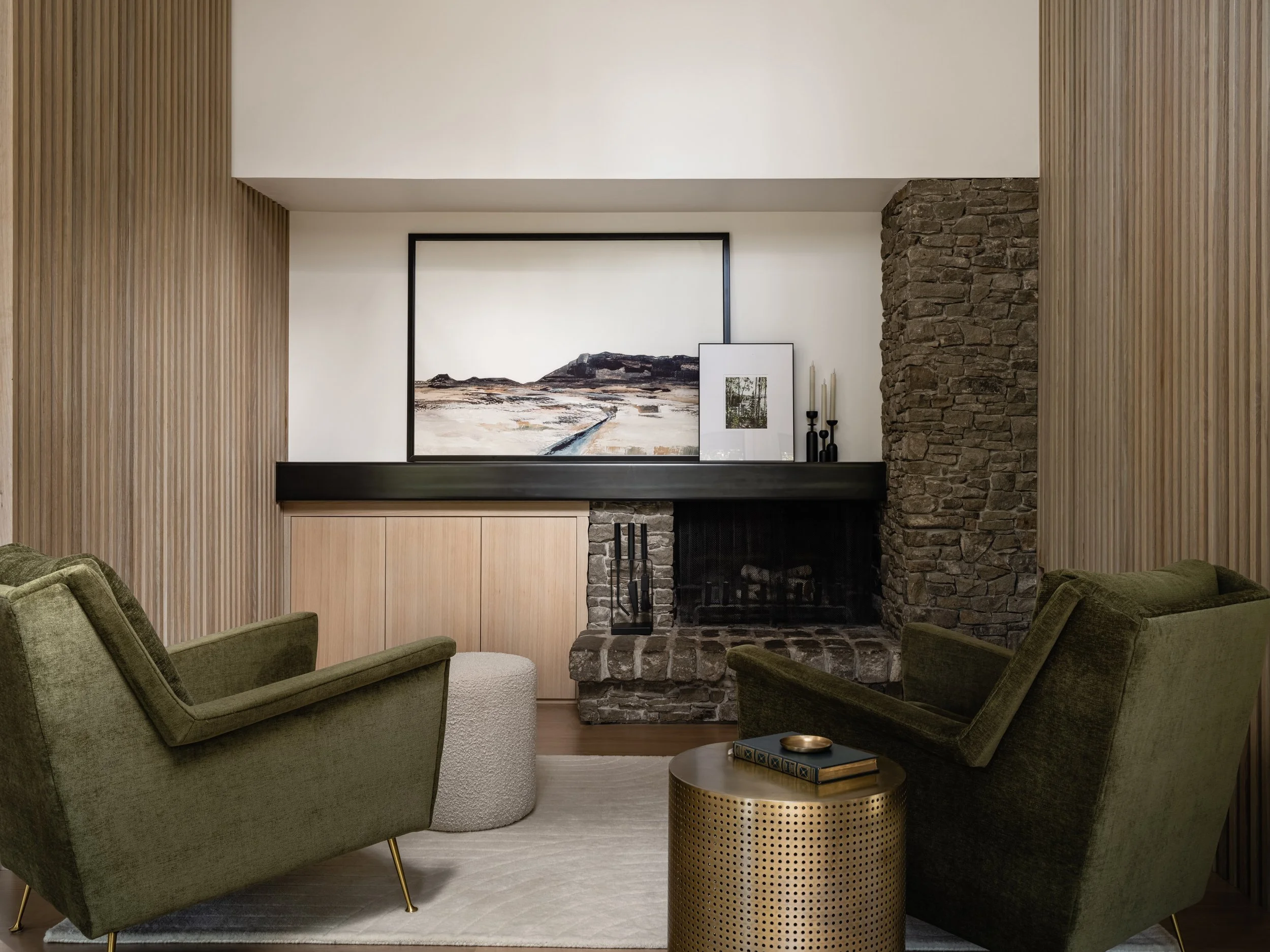 Modern living room featuring green armchairs, a textured stone fireplace, framed wall art above, wood paneling, and a round metallic side table.