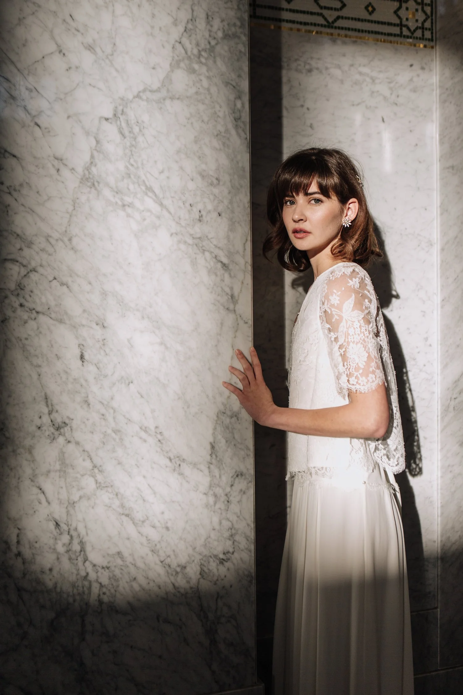 Woman in white lace dress standing by marble wall.