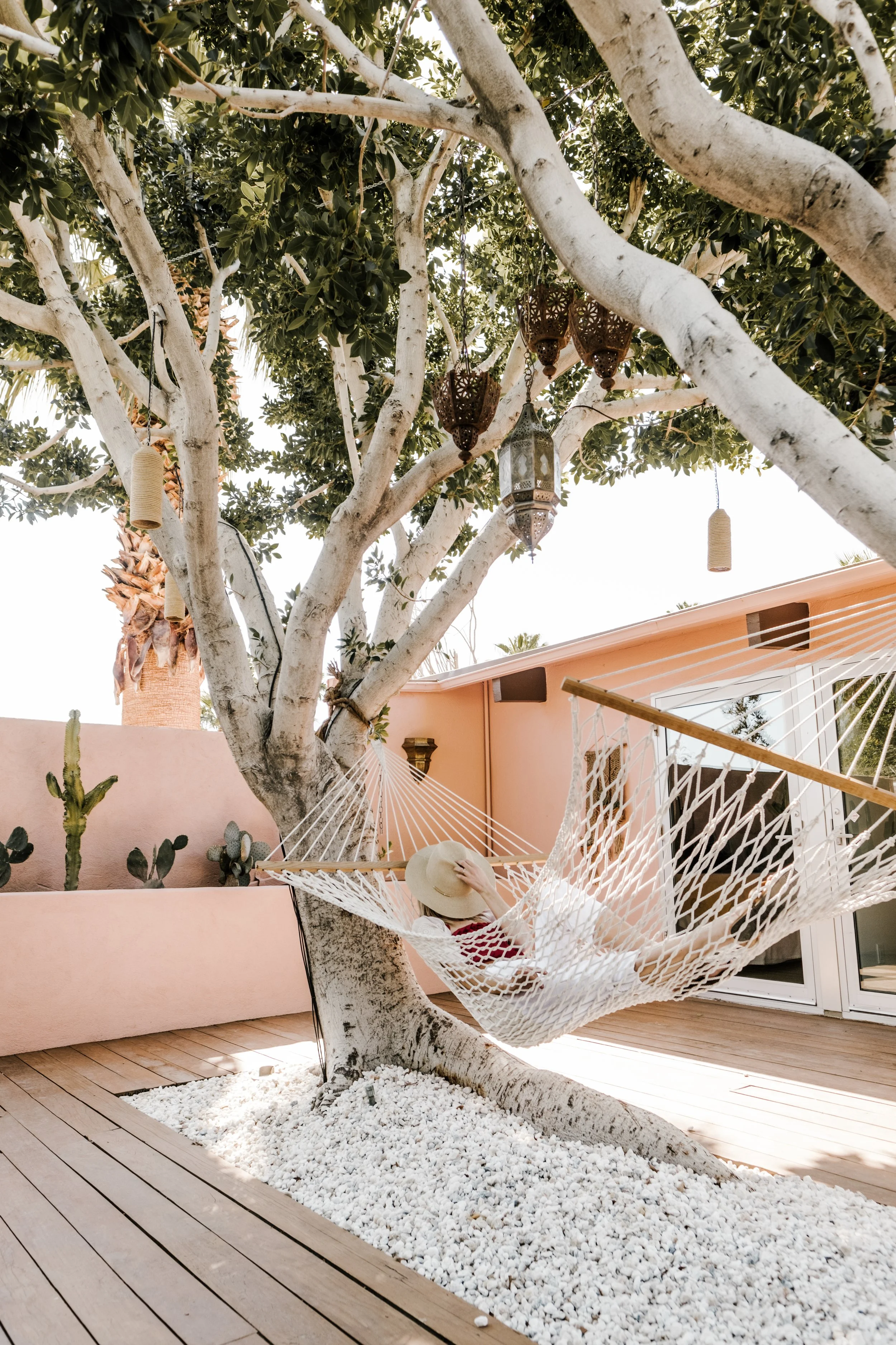 A person relaxing in a hammock under a tree on a wooden deck. The tree is surrounded by white stones, and there are decorative lanterns hanging from its branches. The background features a peach-colored wall with cacti.