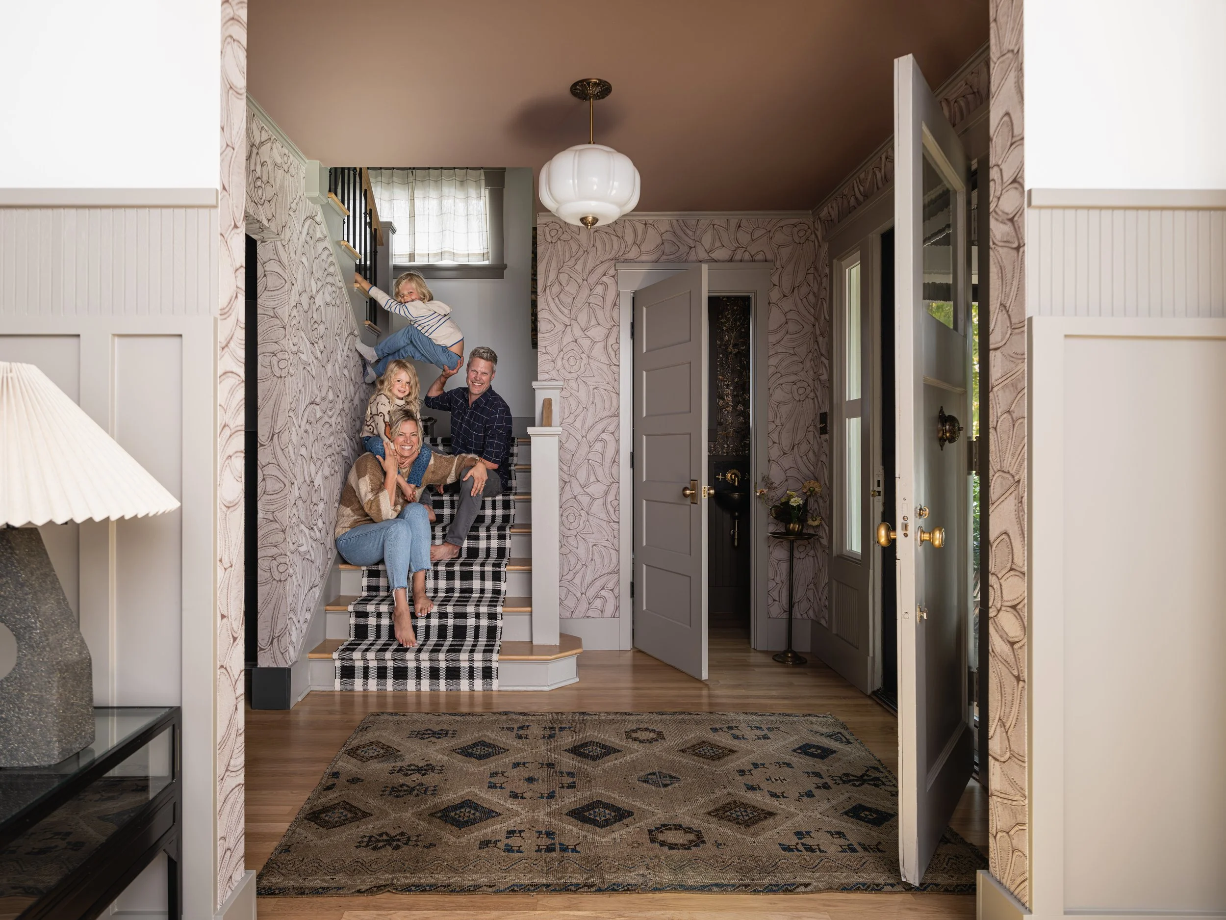 Family sitting on staircase in a warm, stylish home entryway. Adults and children smile, with floral wallpaper and a checkered stair runner. Open front door and a patterned rug on hardwood floor.