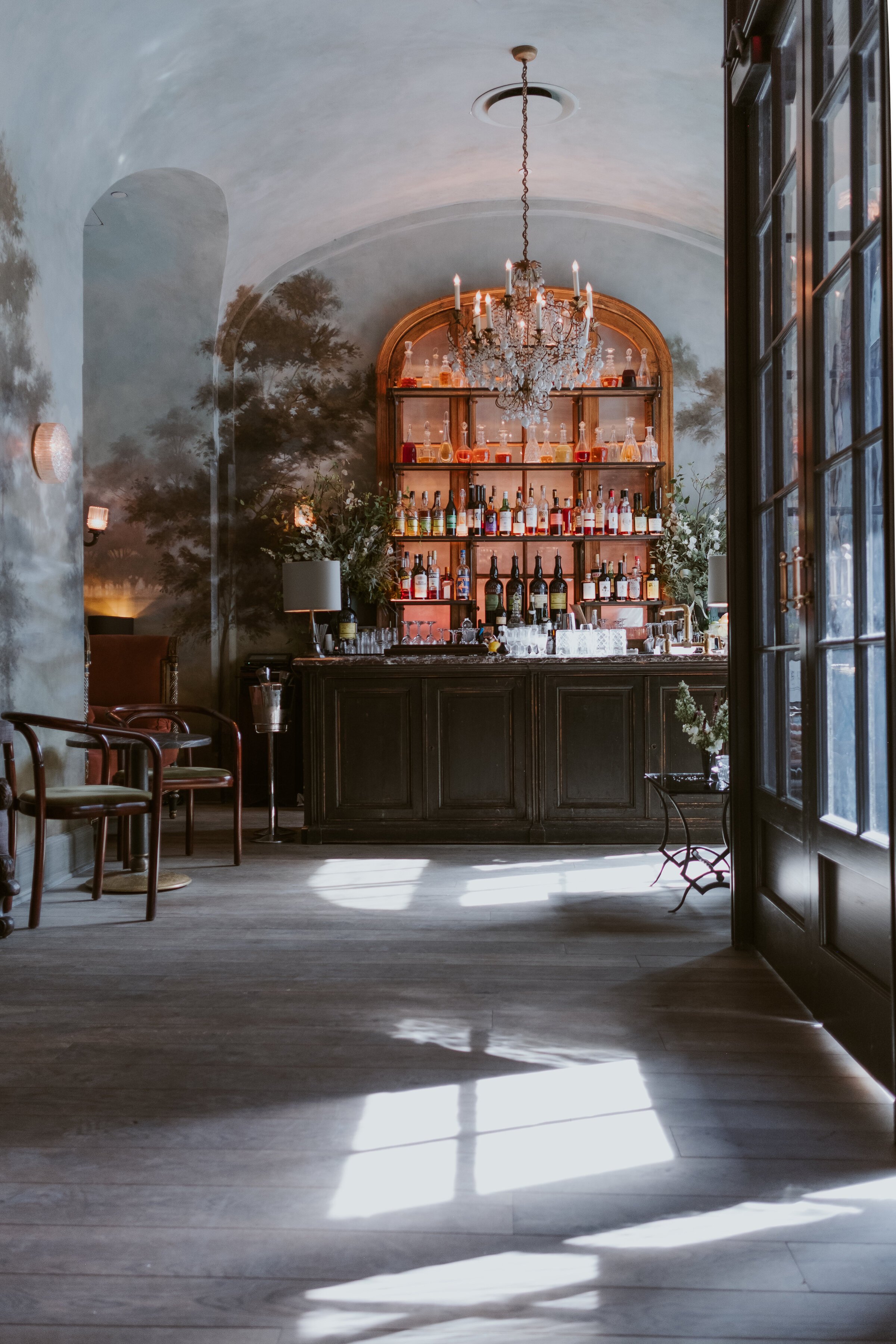 Elegant bar interior with a chandelier, decorative wall art, and various bottles on shelves behind a wooden counter.