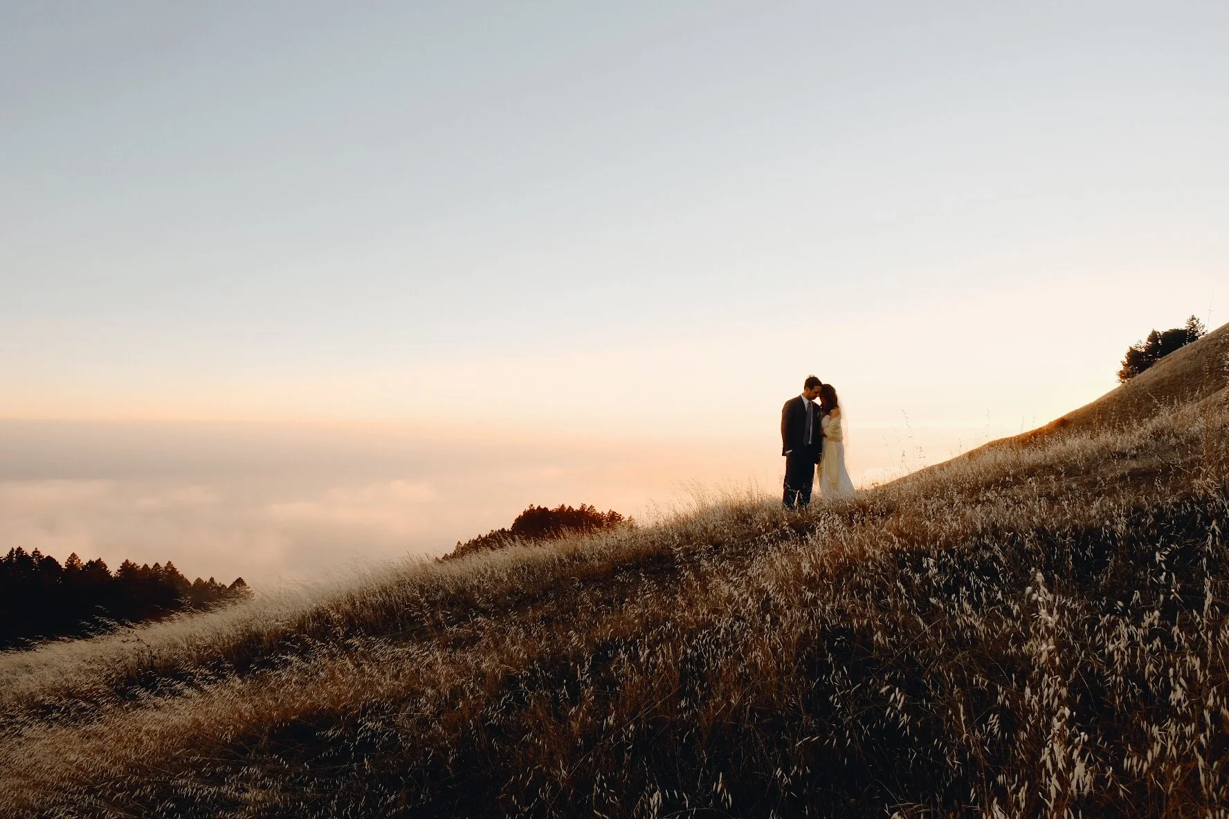 A couple stands on a grassy hill at sunset, embracing under a clear sky.