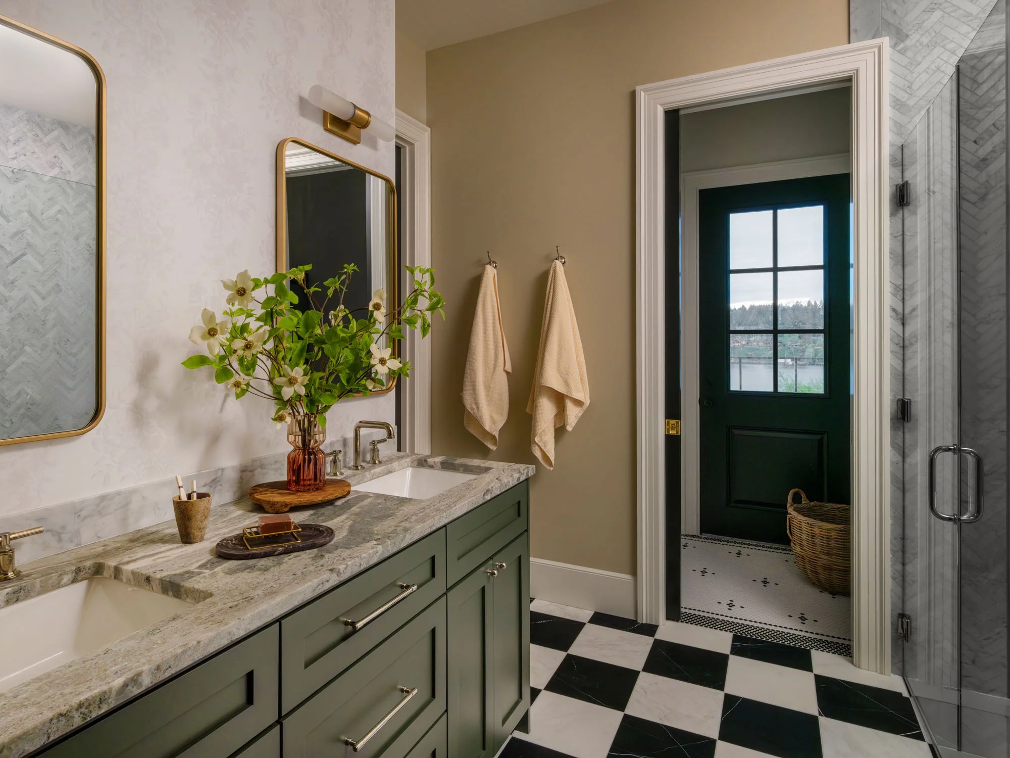 A bathroom with a green vanity, marble countertop, and two mirrors. A vase with flowers is on the counter. Two towels hang on the wall. There is a door leading outside with a window, and part of a shower with marble tiles.
