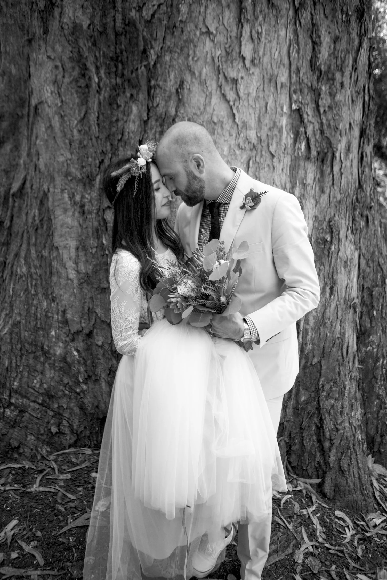 Black and white photo of a bride and groom embracing in front of a large tree trunk, with the bride holding a bouquet.