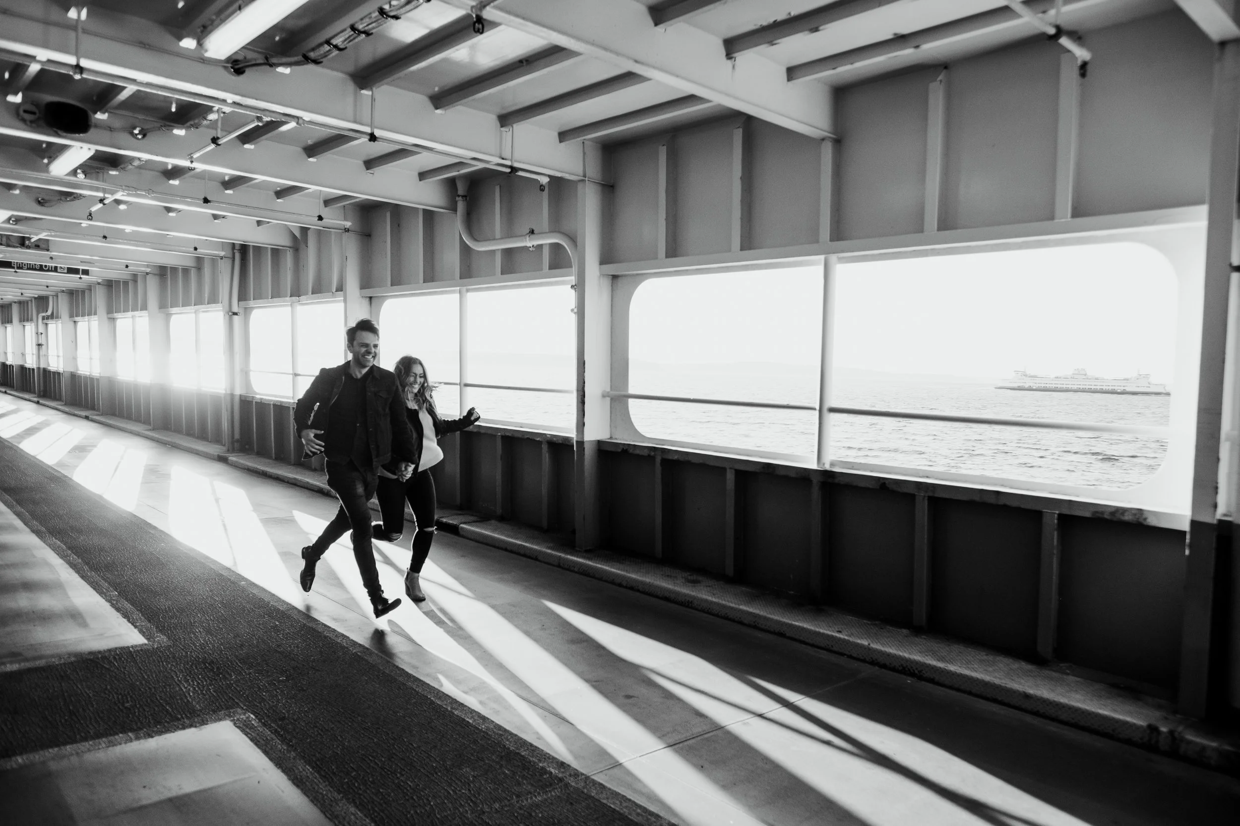 Couple holding hands running on a ferry deck with water visible through windows.