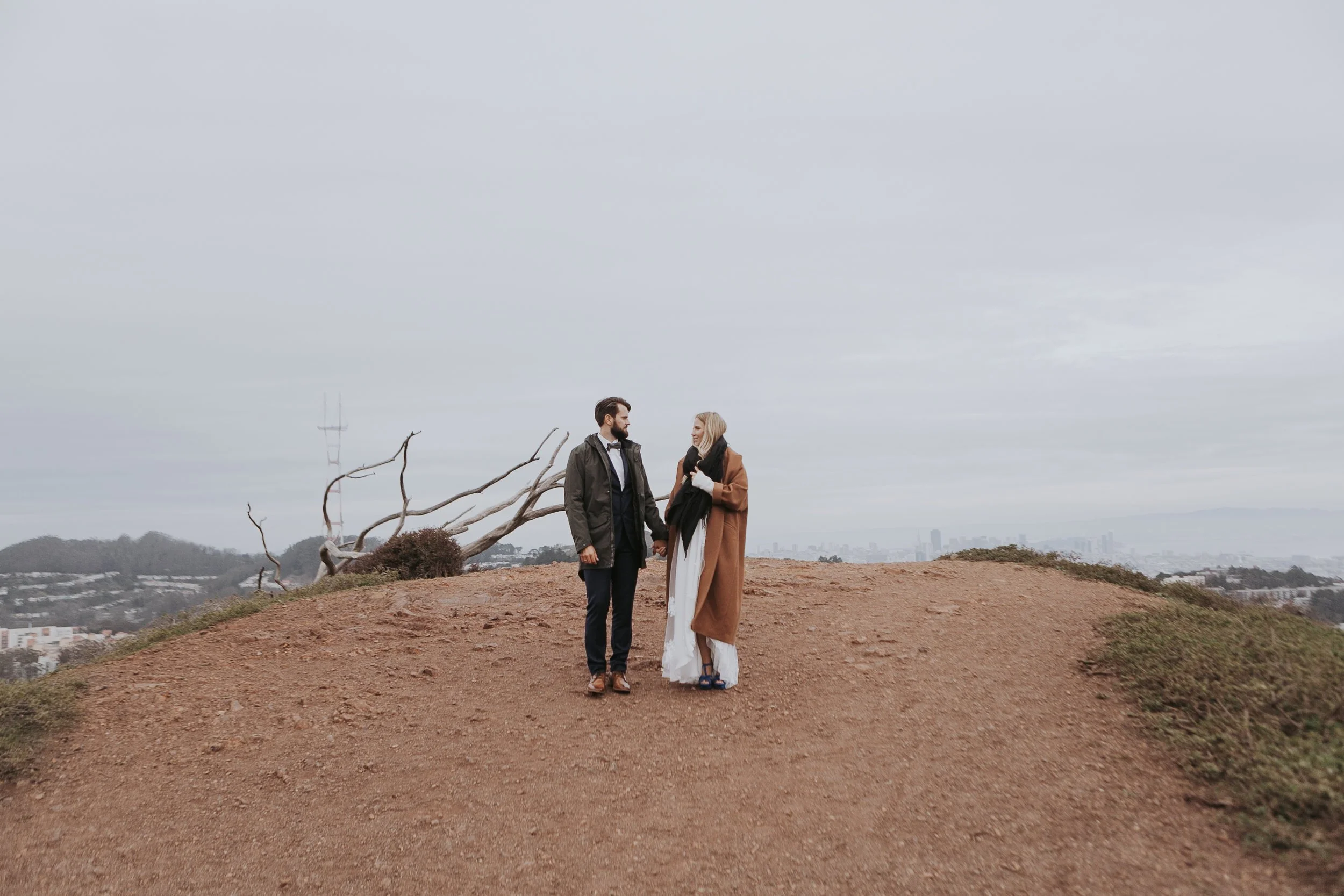 A couple standing on a hill with a cityscape in the background, holding hands and looking at each other. The sky is overcast and there is a bare tree nearby.