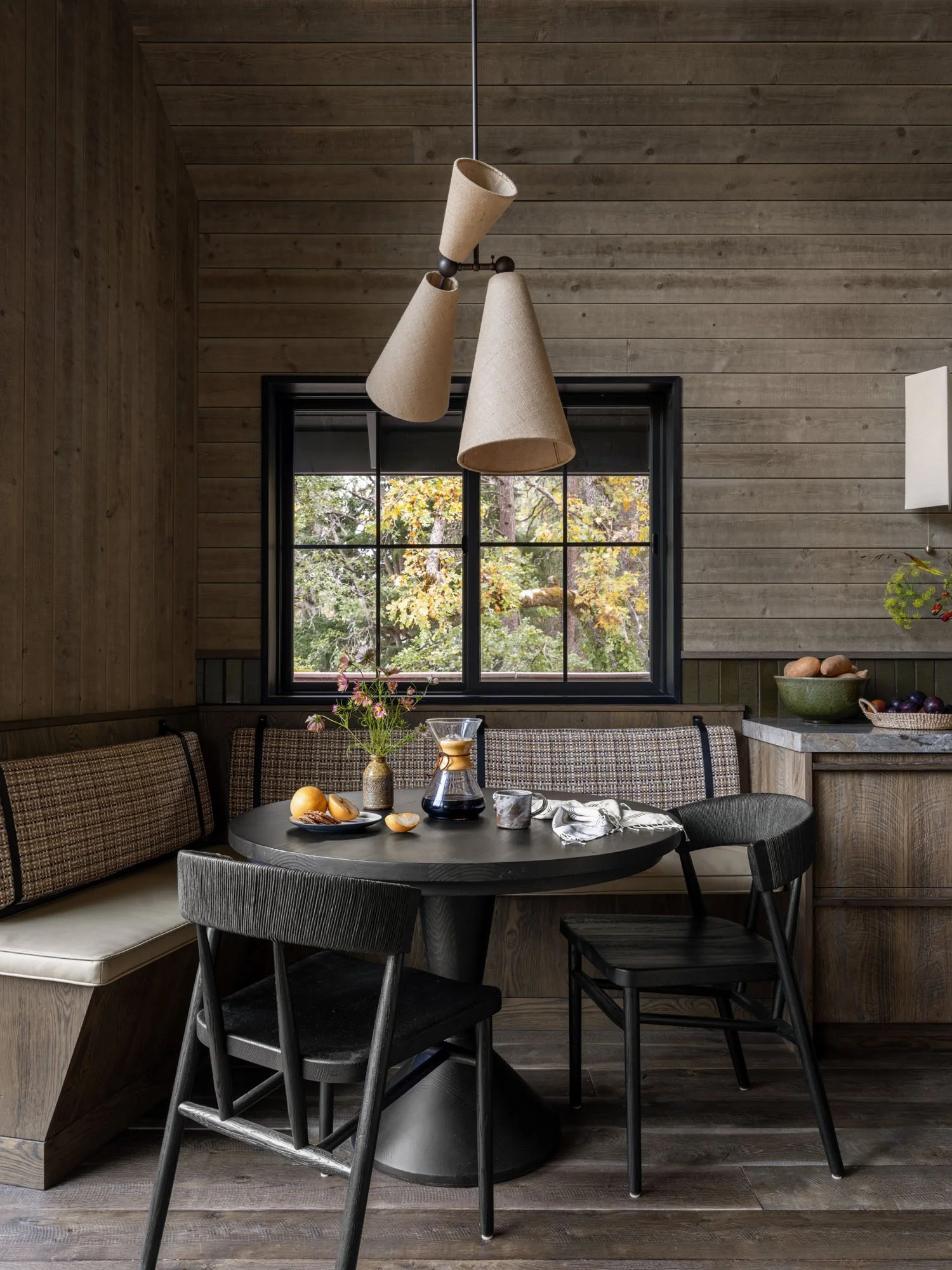 Rustic dining nook with a black round table, black and plaid chairs, a window with trees outside, and a pendant light fixture