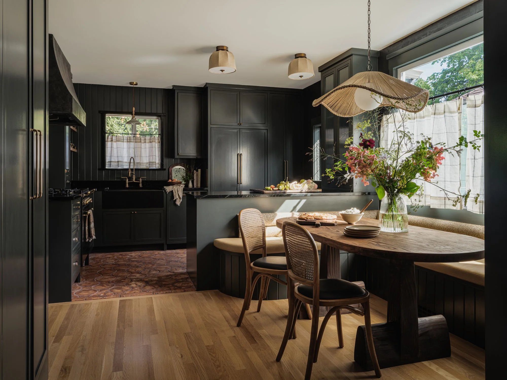 A kitchen with black cabinets, a black sink, and a dark wood dining table with chairs and a bench. A flower arrangement in a glass vase is on the table near a window with white curtains. Light wood floor and ceiling lighting fixtures complete the sce