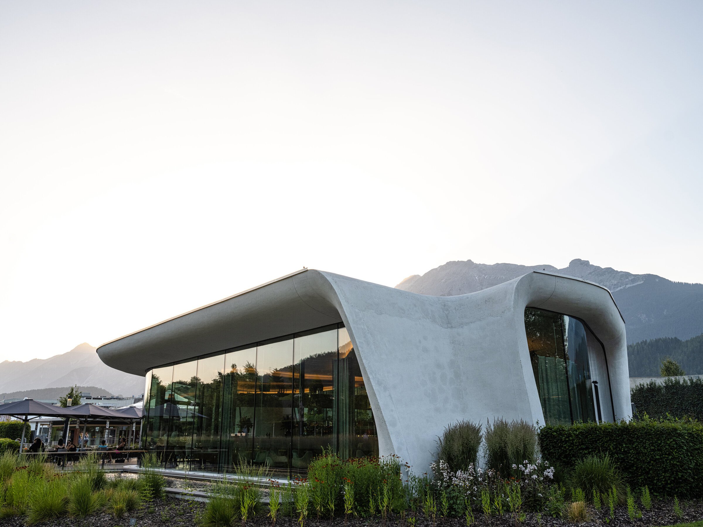 Futuristic building with curved architecture and glass walls, surrounded by landscaped gardens, with mountains in the background.
