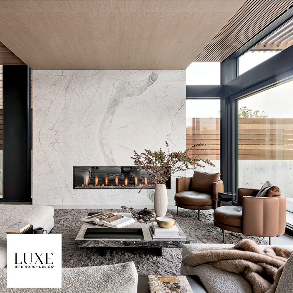 Modern living room with a white marble wall, horizontal fireplace, large windows with black frames, tan leather chairs, a white sofa, a black coffee table with books and decorative items, a gray rug, a potted plant, and a blanket on the sofa.