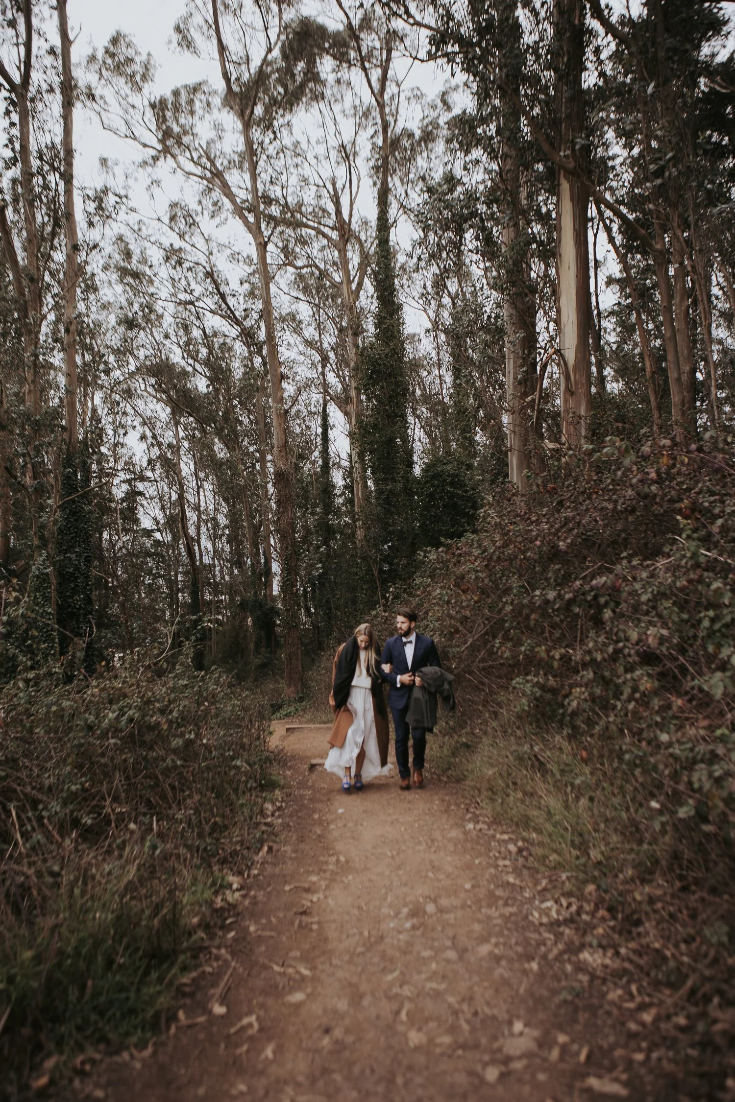 Couple walking on a forest path surrounded by tall trees.