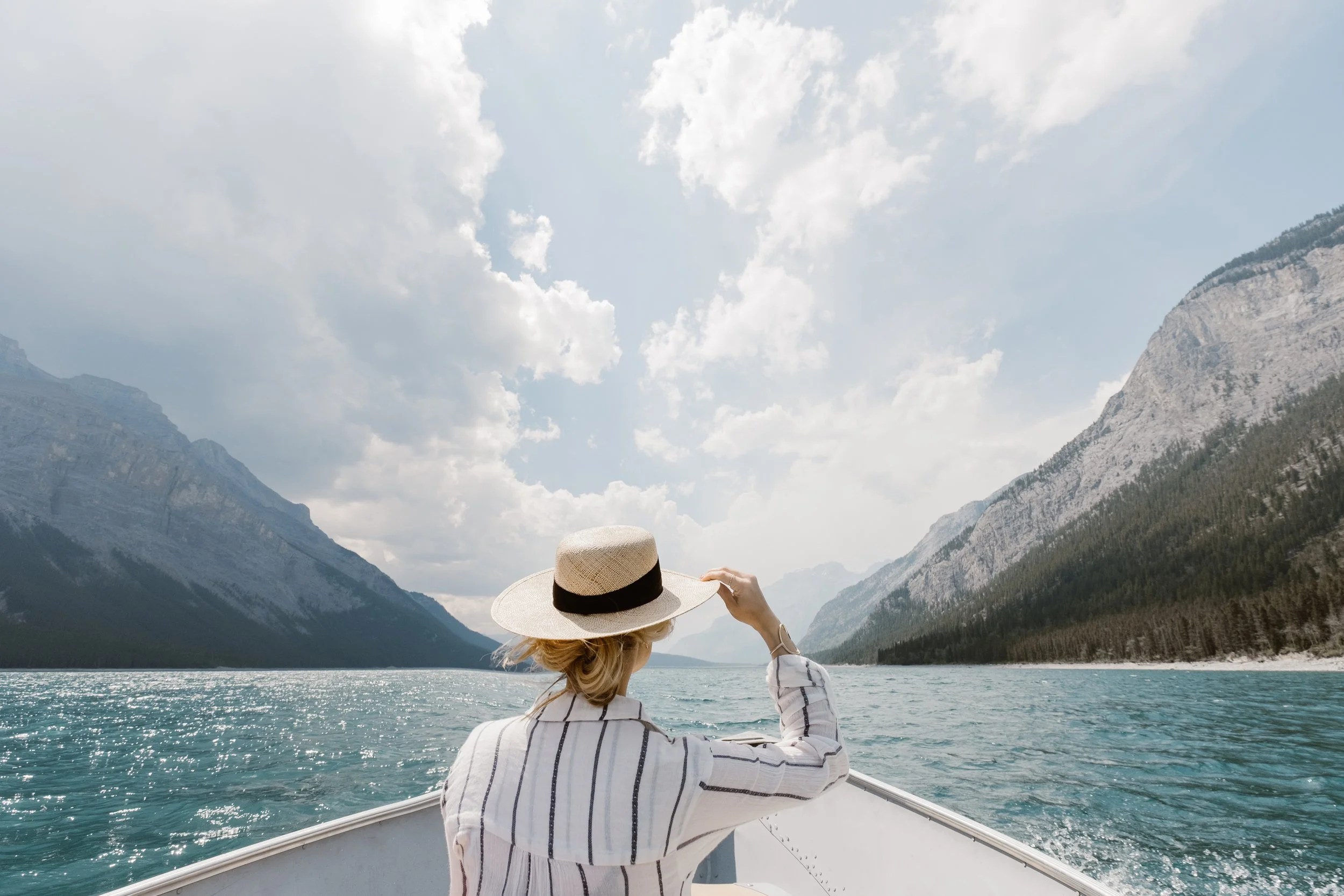 Person wearing a hat on a boat, surrounded by a lake and mountains under a cloudy sky.