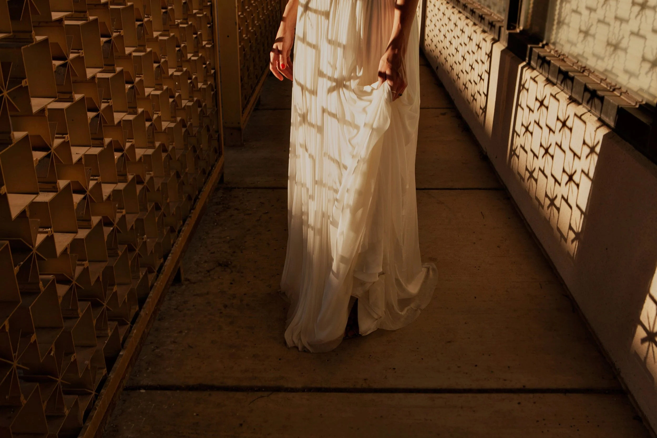 Person holding the bottom of a long white dress in a corridor, with geometric shadows on the walls.