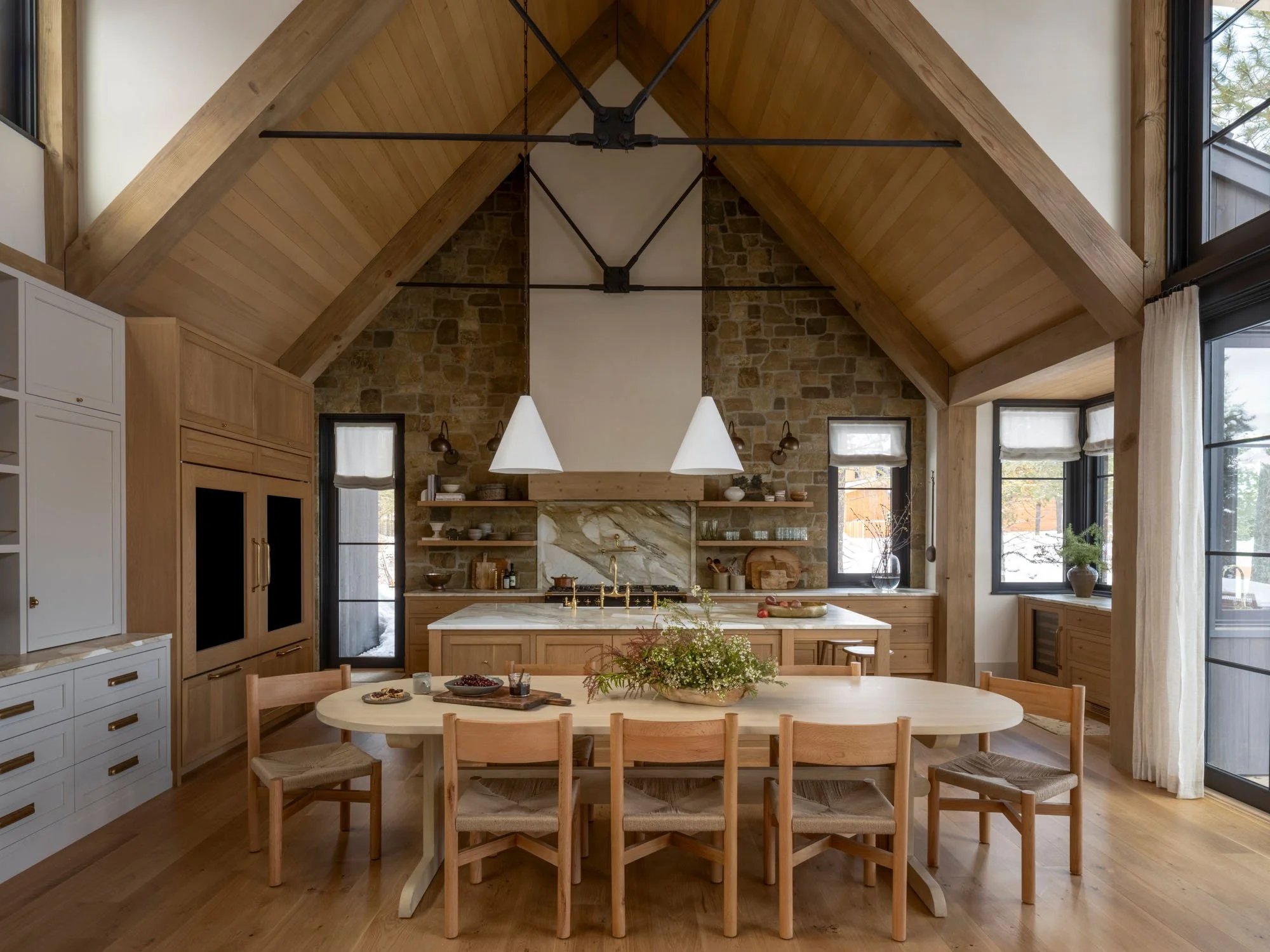 A rustic kitchen with a high, vaulted wooden ceiling, exposed brick wall behind the stove, and large windows. There is an oval dining table with eight chairs, pendant lights above, wooden cabinetry, and a mix of modern and vintage decor.