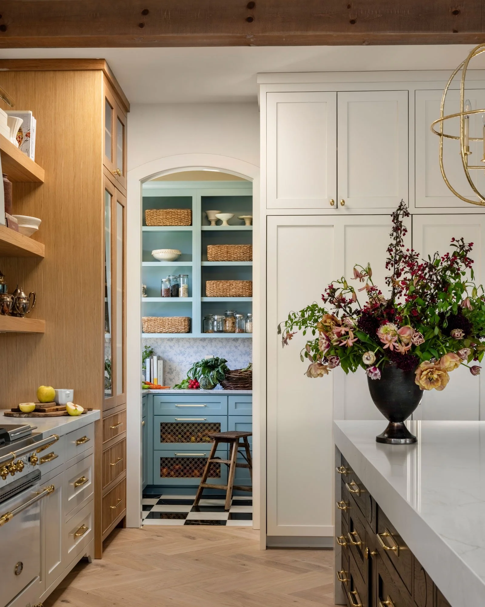 A well-organized kitchen with white and natural wood cabinetry, a white marble countertop, and a large floral arrangement on the island. A view into a small but colorful adjacent kitchen or pantry space is visible through an open archway.