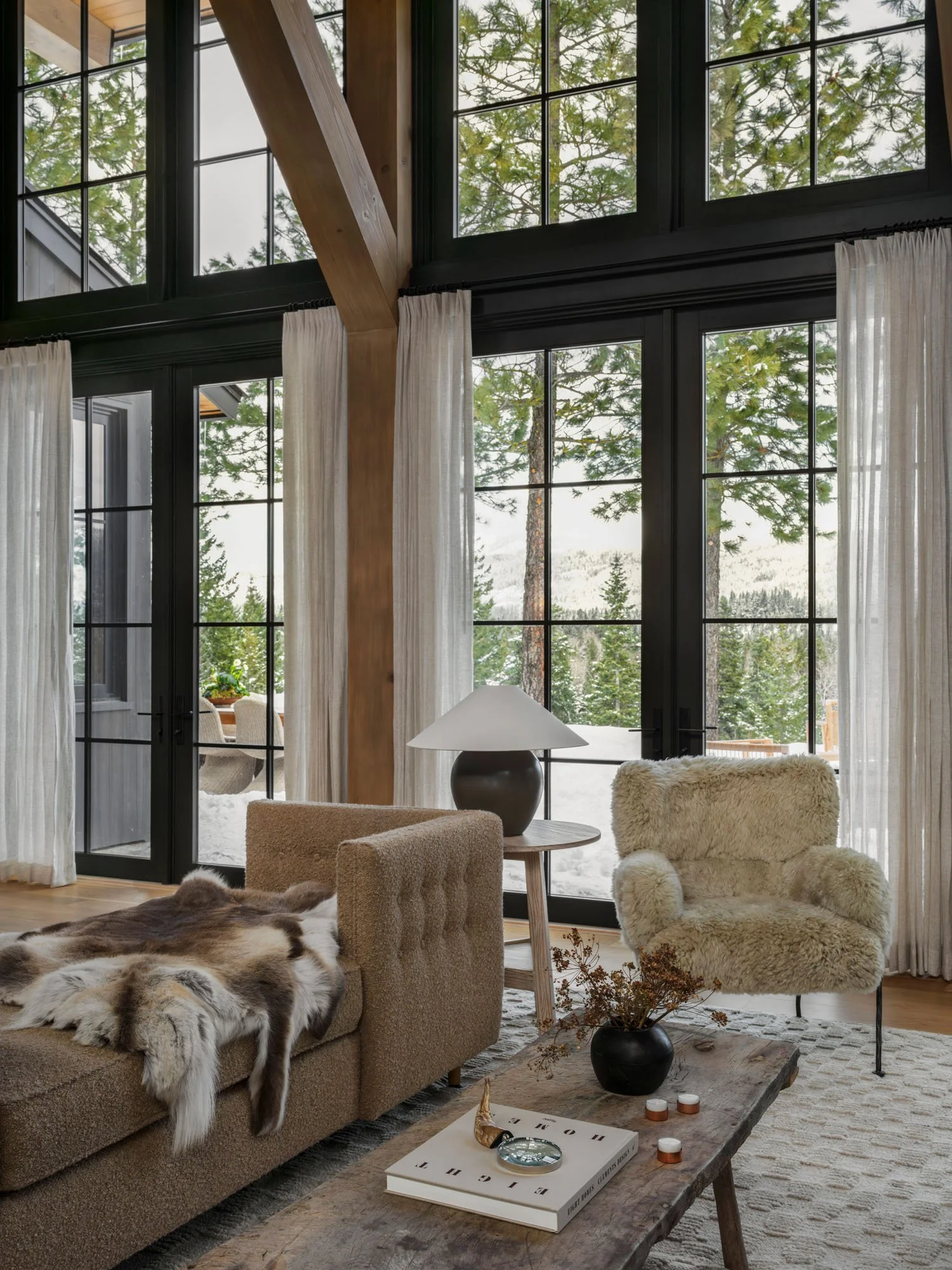 Living room with large windows, beige couch with a fur blanket, plush chair, wooden coffee table with a black vase and candles, and a lamp, overlooking a snowy landscape with trees.