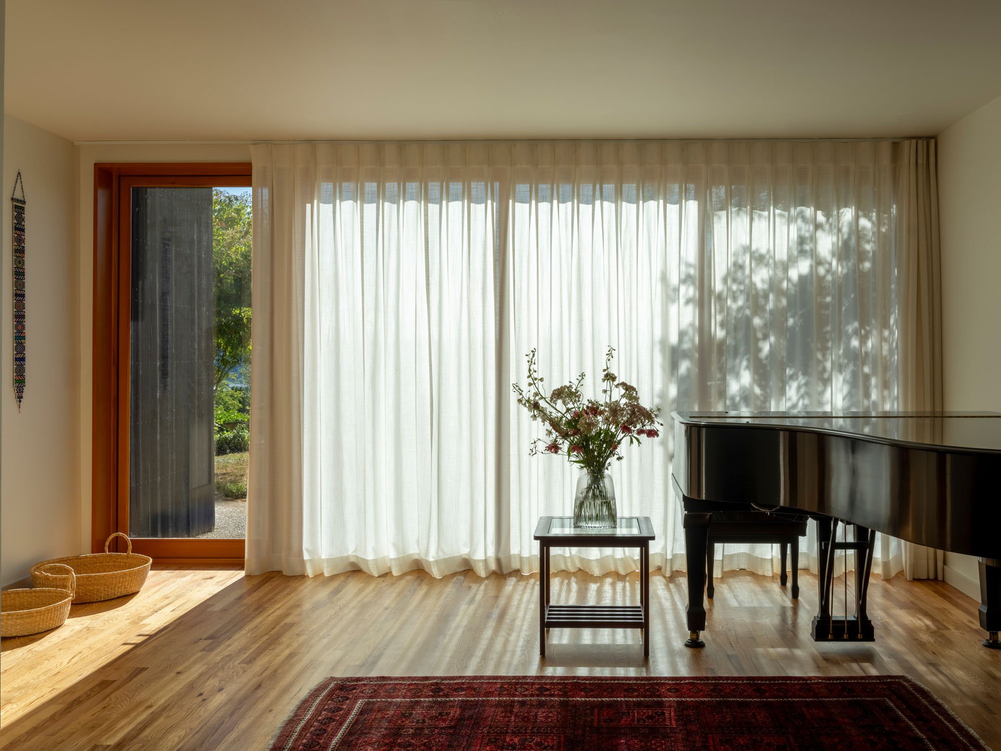 Bright living room with large windows covered by sheer white curtains, a black grand piano, a small table with a vase of flowers, and wicker baskets on the wooden floor, with a view of greenery outside.