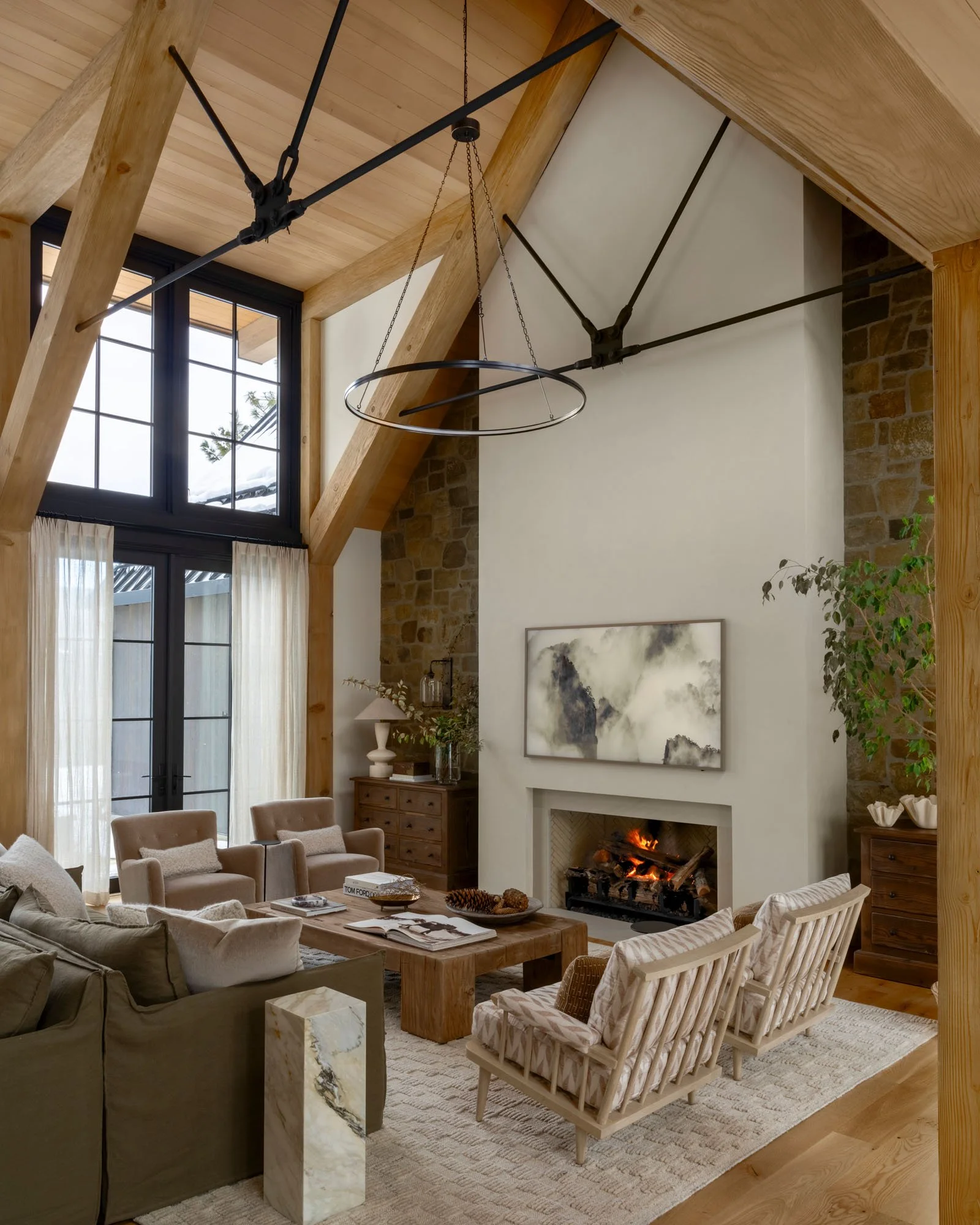 Living room with high ceiling, wood beams, large windows, white walls, a fireplace with a fire, and modern furniture including beige armchairs, a green sofa, and a wooden coffee table.