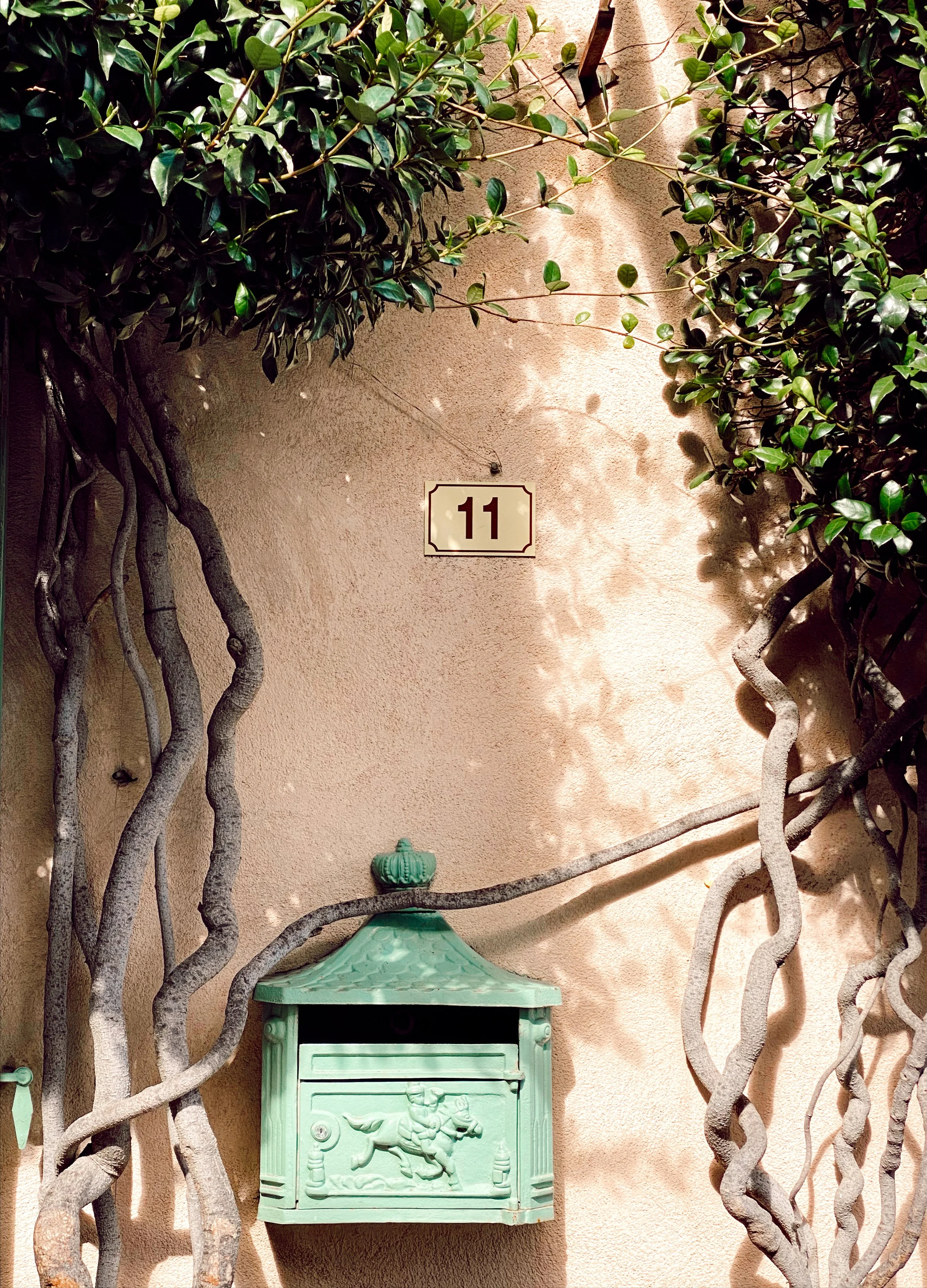 Green vintage mailbox with decorative design on a textured wall, surrounded by leafy vines, and a house number "11" sign above it.