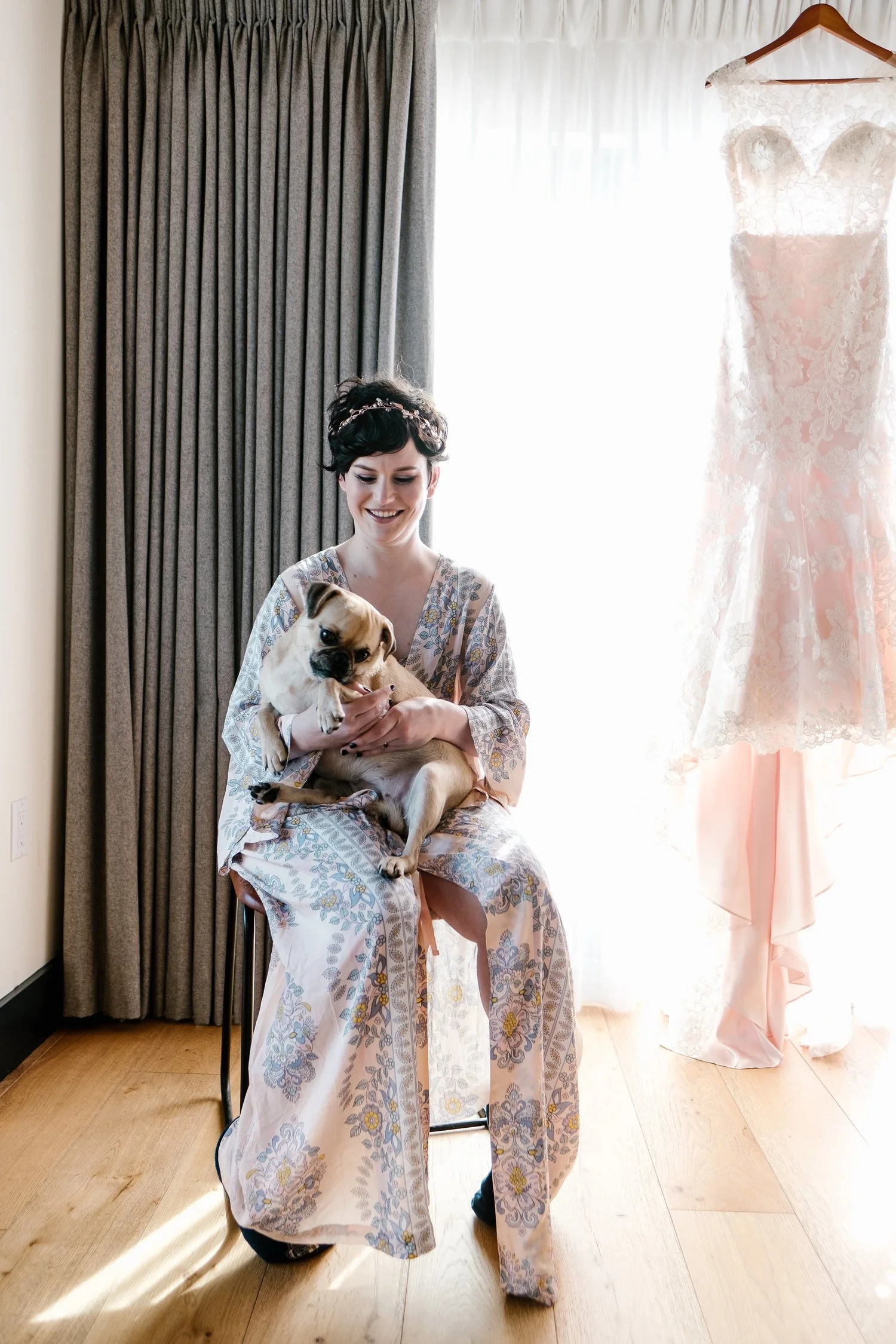 A woman in a floral robe sitting on a stool holding a small dog, with a lace wedding dress hanging nearby, in a softly lit room.