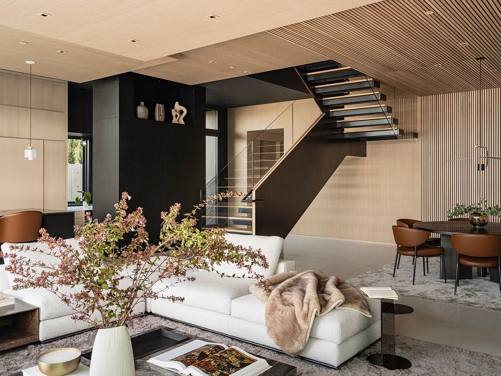 Modern living room with minimalist design, featuring a white sofa, vase with branches, fur throw, open staircase, dining table with brown chairs, and neutral-toned decor.
