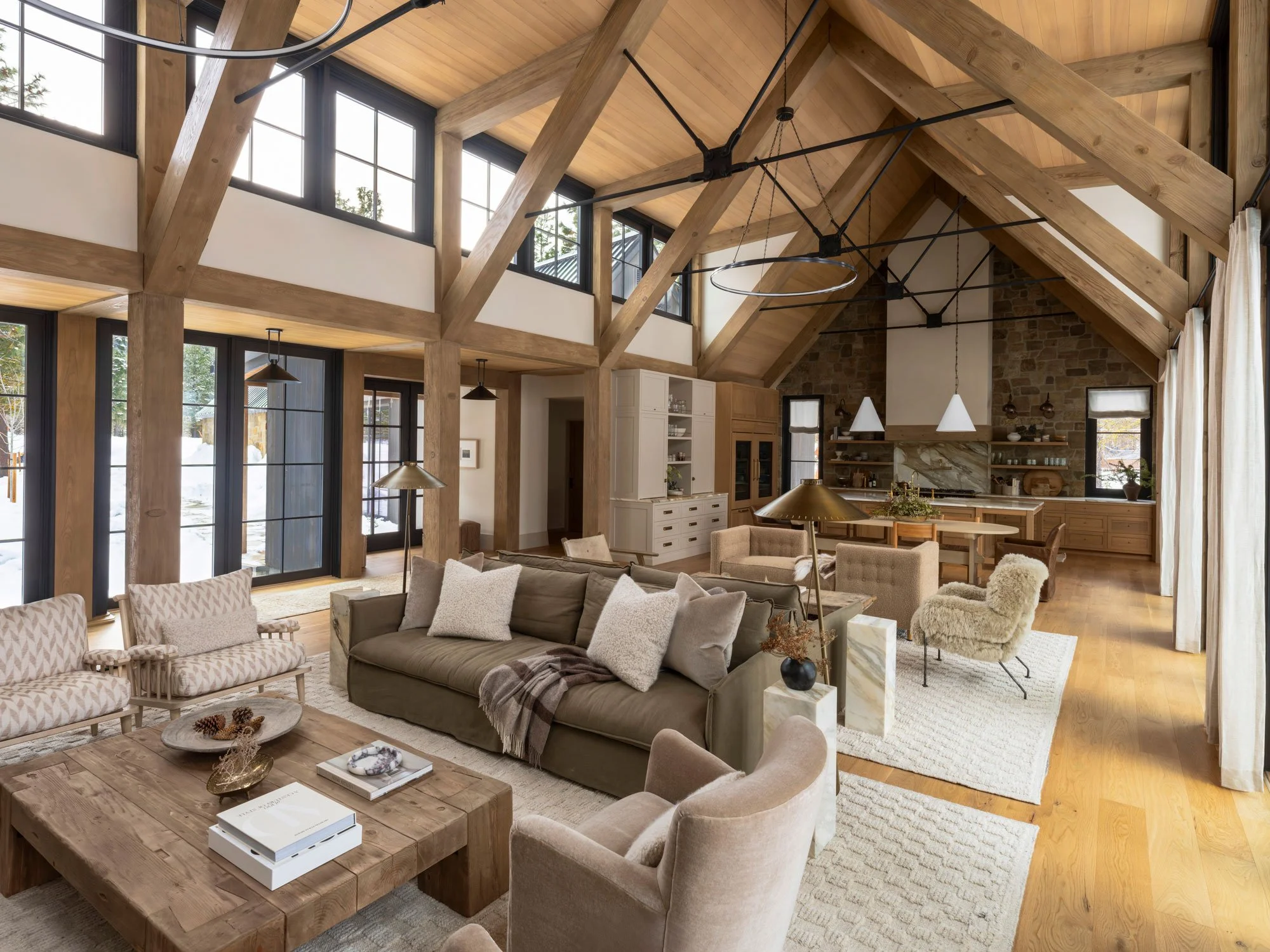 Open-concept living room and kitchen with exposed wooden beams, large windows, and neutral-colored furniture.