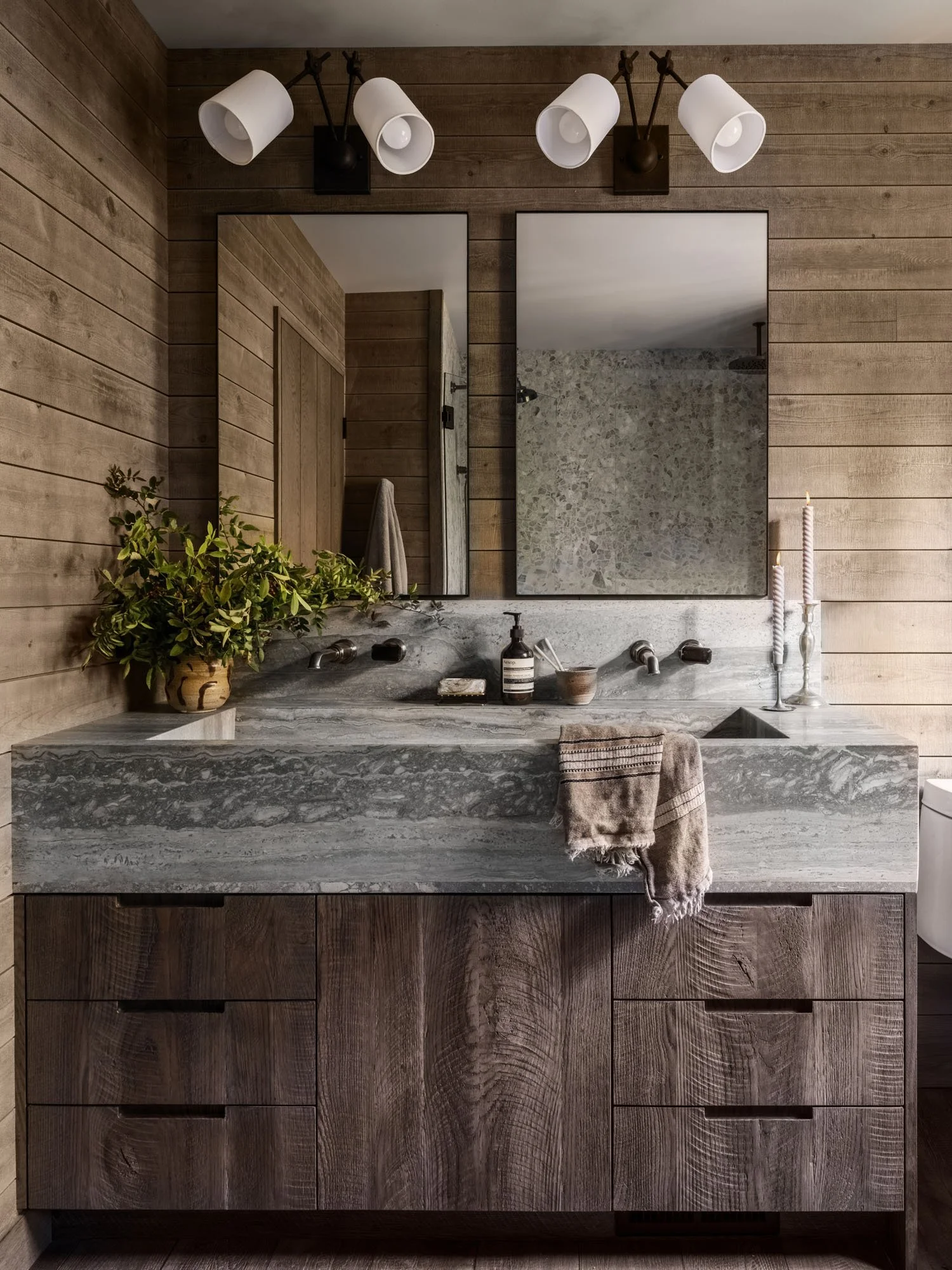 Modern bathroom vanity with a gray marble countertop, wooden cabinet, large mirror, potted green plant, soap dispenser, bowls, two candles, and a towel hanging on the front of the cabinet. The wall behind is wood-paneled and there's a wall-mounted li
