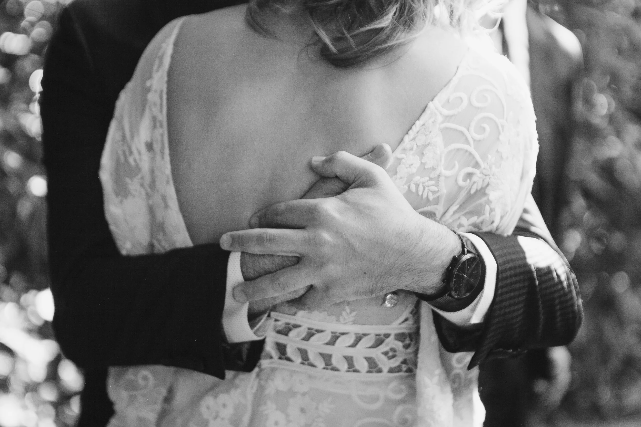 Close-up of a couple embracing, with one wearing a lace dress and the other in a suit. Focus on hands and garment details.