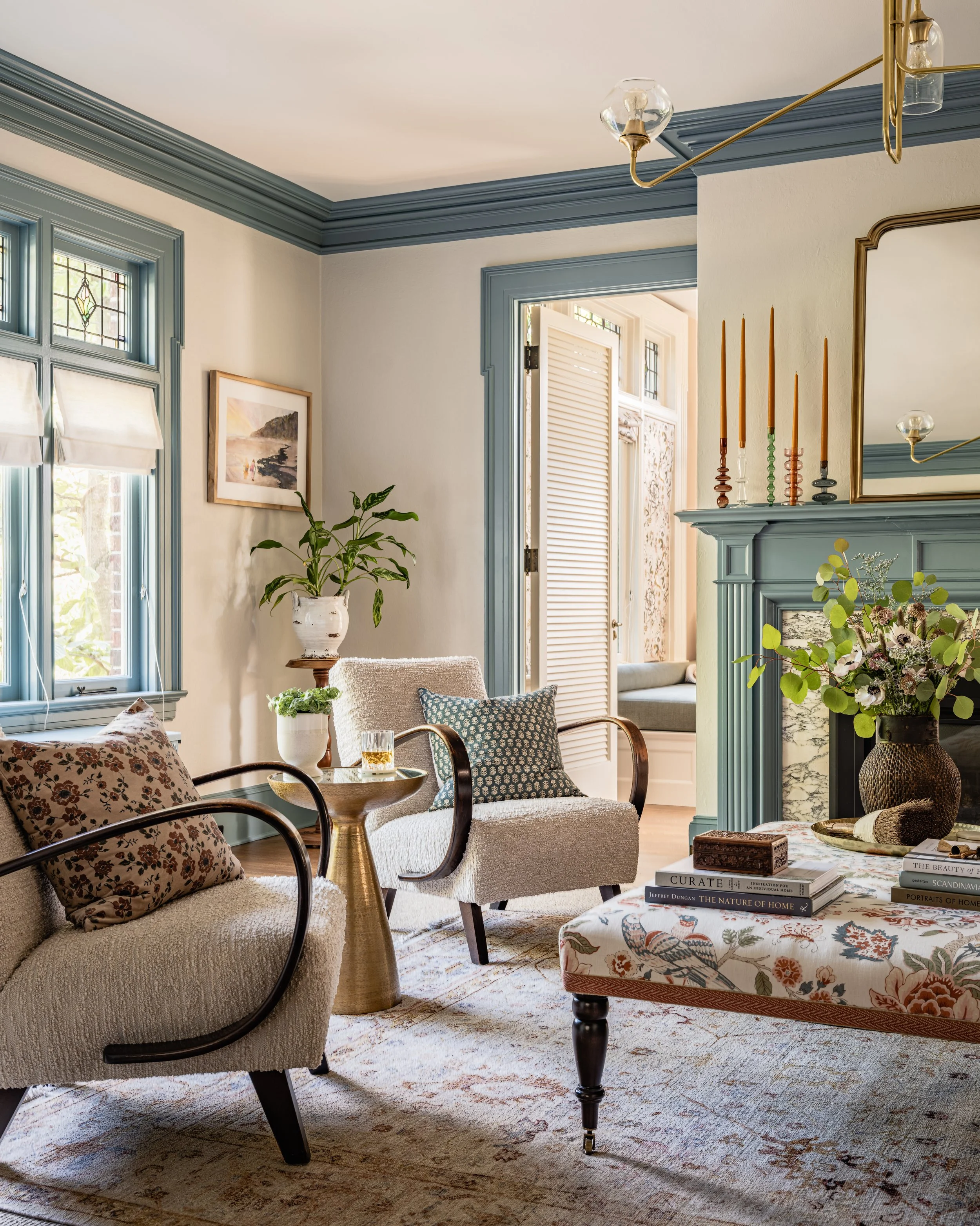 A cozy living room with two cushioned armchairs and a small round side table between them. The decor includes a floral-patterned bench, a fireplace mantel with tall candlesticks, and a framed picture beside a window. A houseplant is placed on a stand