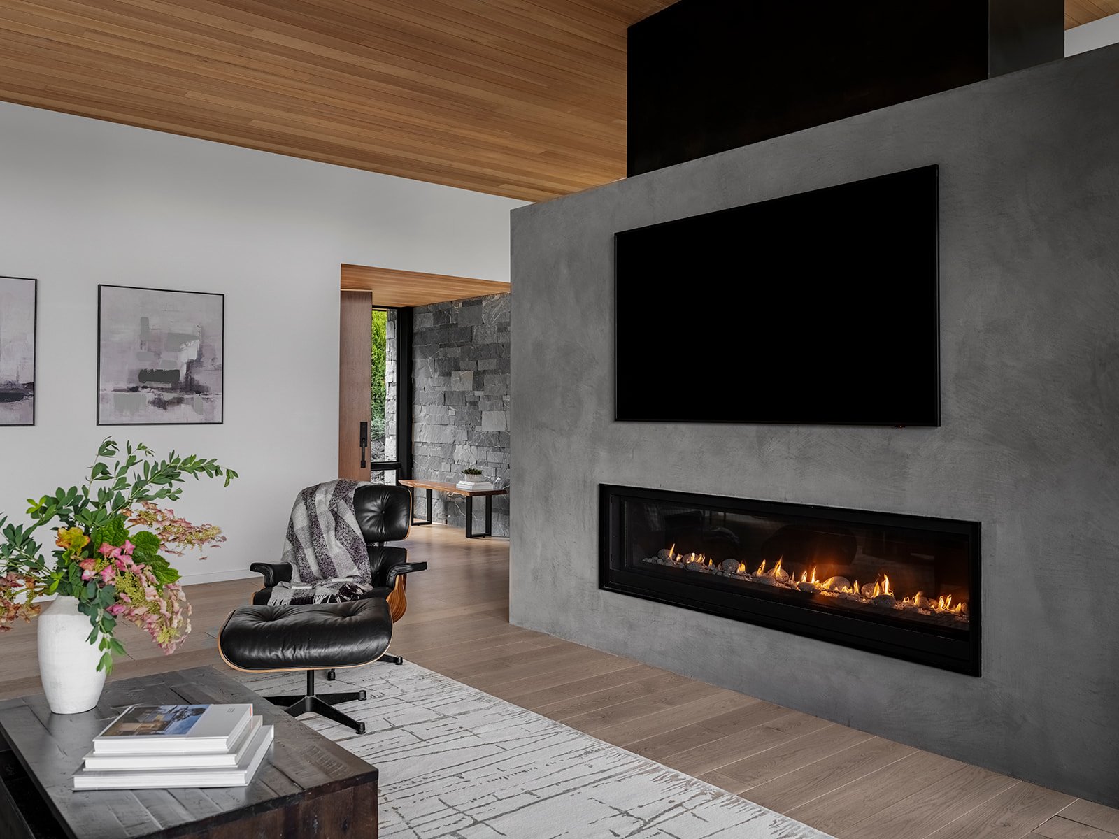 Modern living room with a fireplace and television, featuring a black leather chair with a blanket, a coffee table with books and flowers, and artwork on the walls.