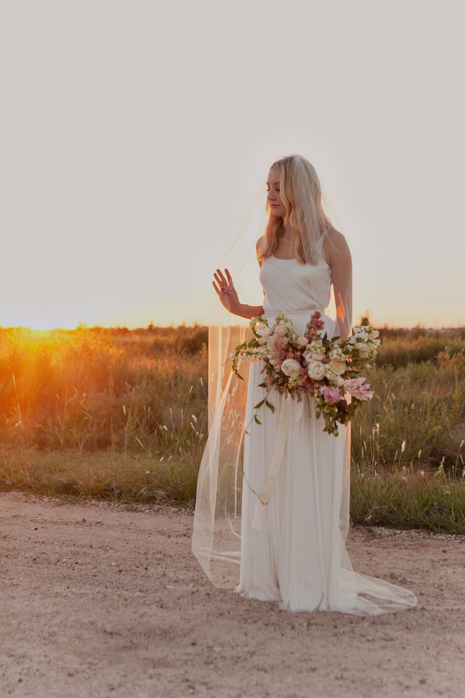 Bride in a white gown and veil holding a bouquet, standing in a field at sunset.
