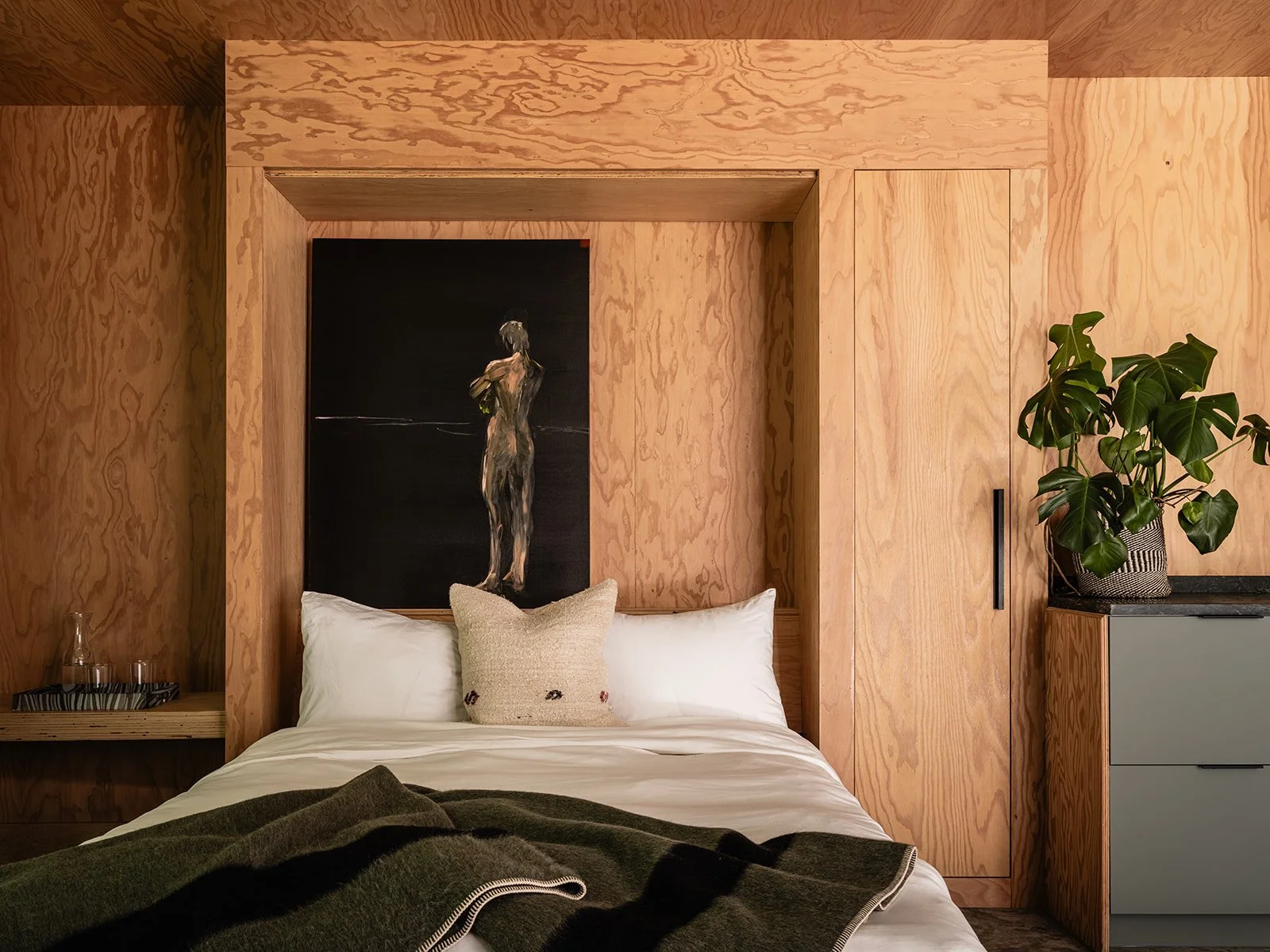 Bedroom with a wooden wall and ceiling, featuring a bed with white bedding, a green blanket, and a textured cushion. A painting of a figure is placed above the bed. A potted plant sits atop a wooden cabinet.