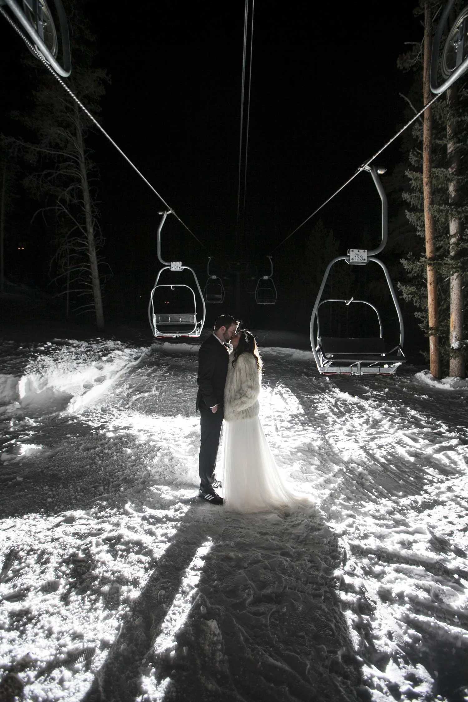 A couple standing on a snowy path under a ski lift at night, with the woman wearing a white gown and fur jacket, and the man in a dark suit.