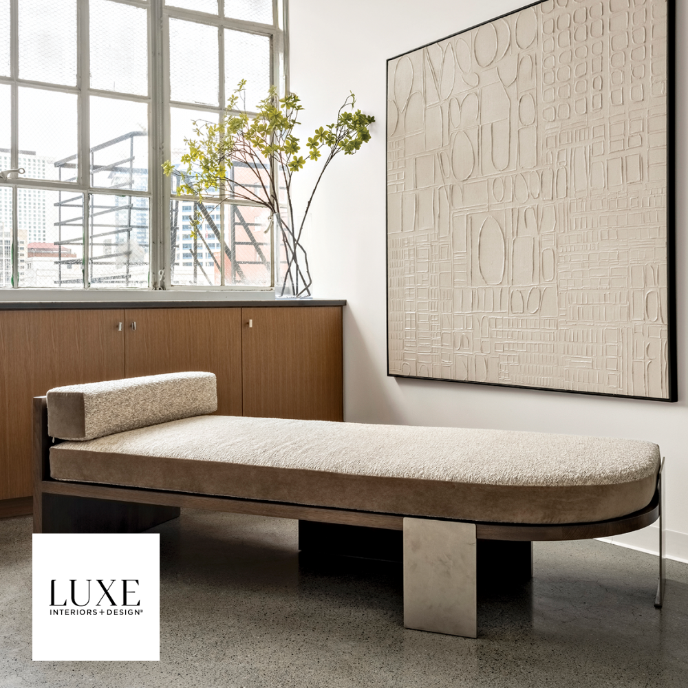 A beige upholstered chaise lounge with a rectangular headrest in a modern, minimalist room. The room features a large, textured abstract artwork on the wall, a wooden cabinet with a plant on top, and large windows with a cityscape view.