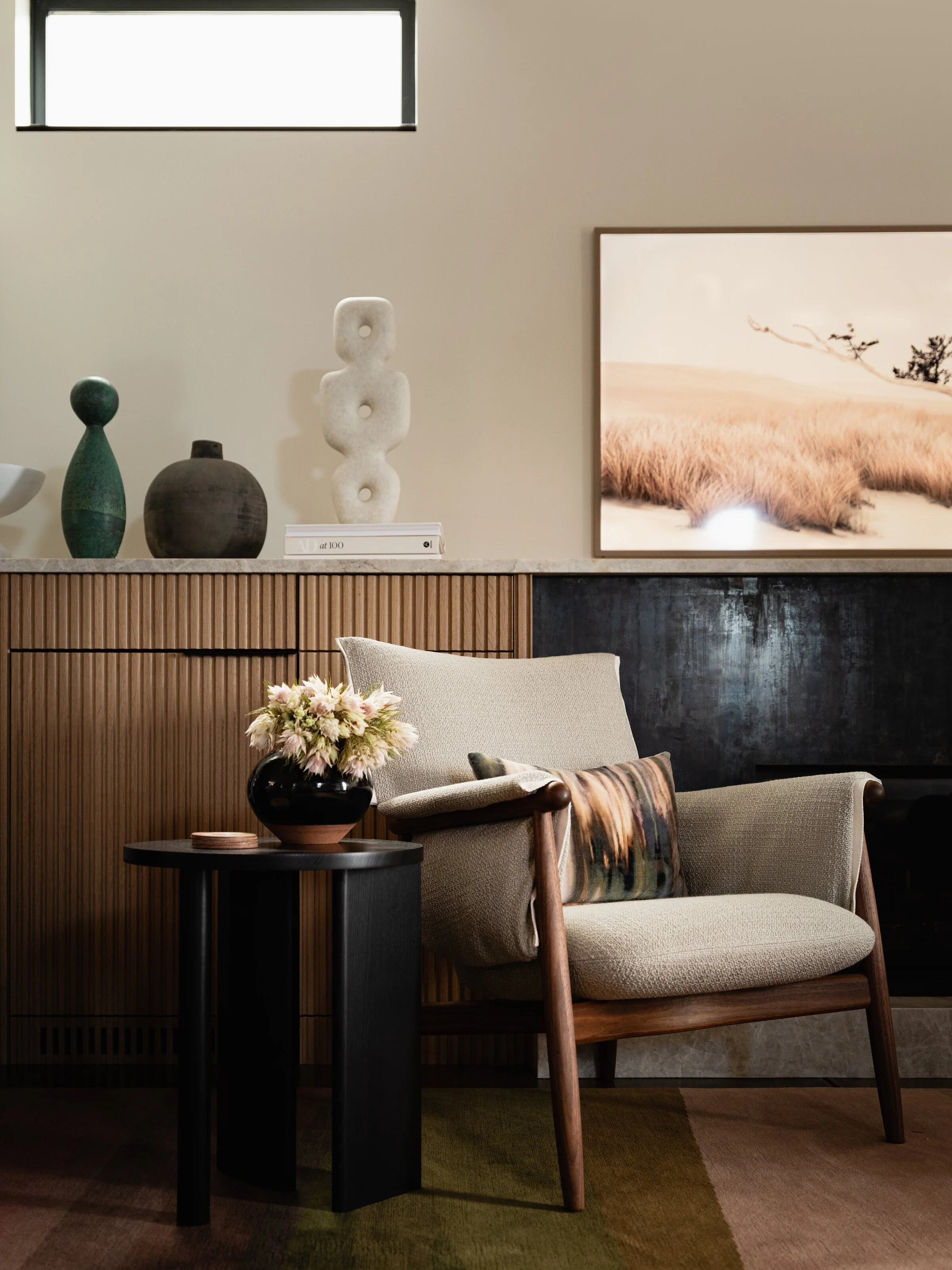 Modern living room with a beige armchair, black side table, vase with flowers, abstract sculpture, and landscape photograph.