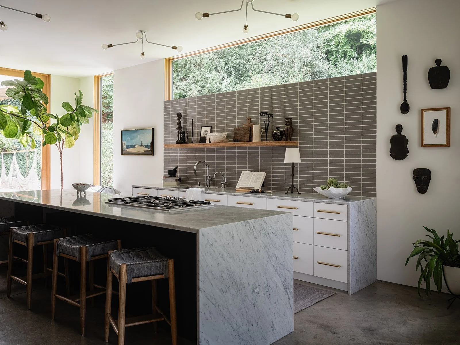 Modern kitchen with large marble island, bar stools, gray tile backsplash, open shelves, decorative wall art, and a leafy plant next to the window.