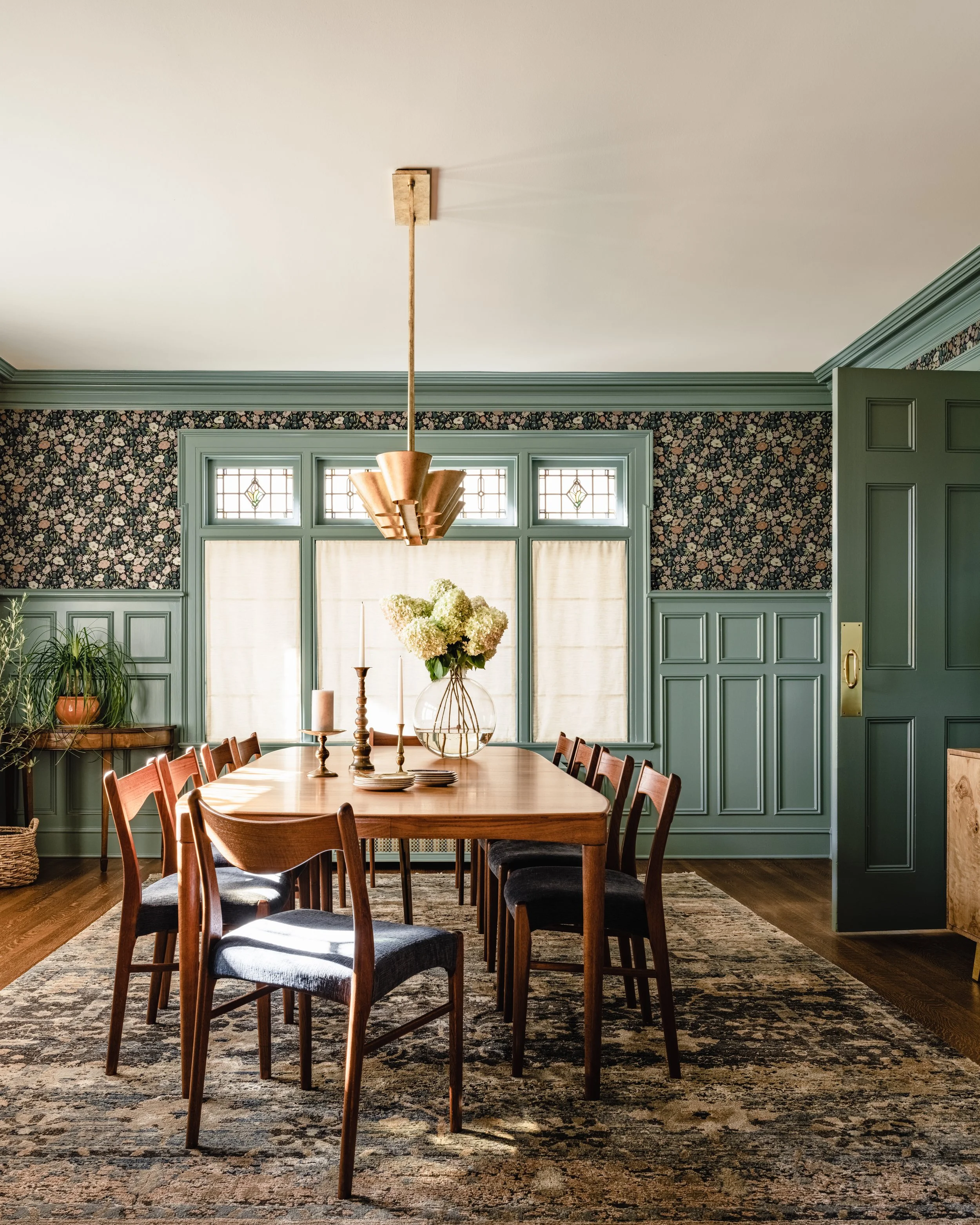 Elegant dining room with a wooden table, eight chairs, floral wallpaper, green paneling, pendant light, and a rug. Decor includes a vase with flowers and candlesticks, with natural light through large windows.