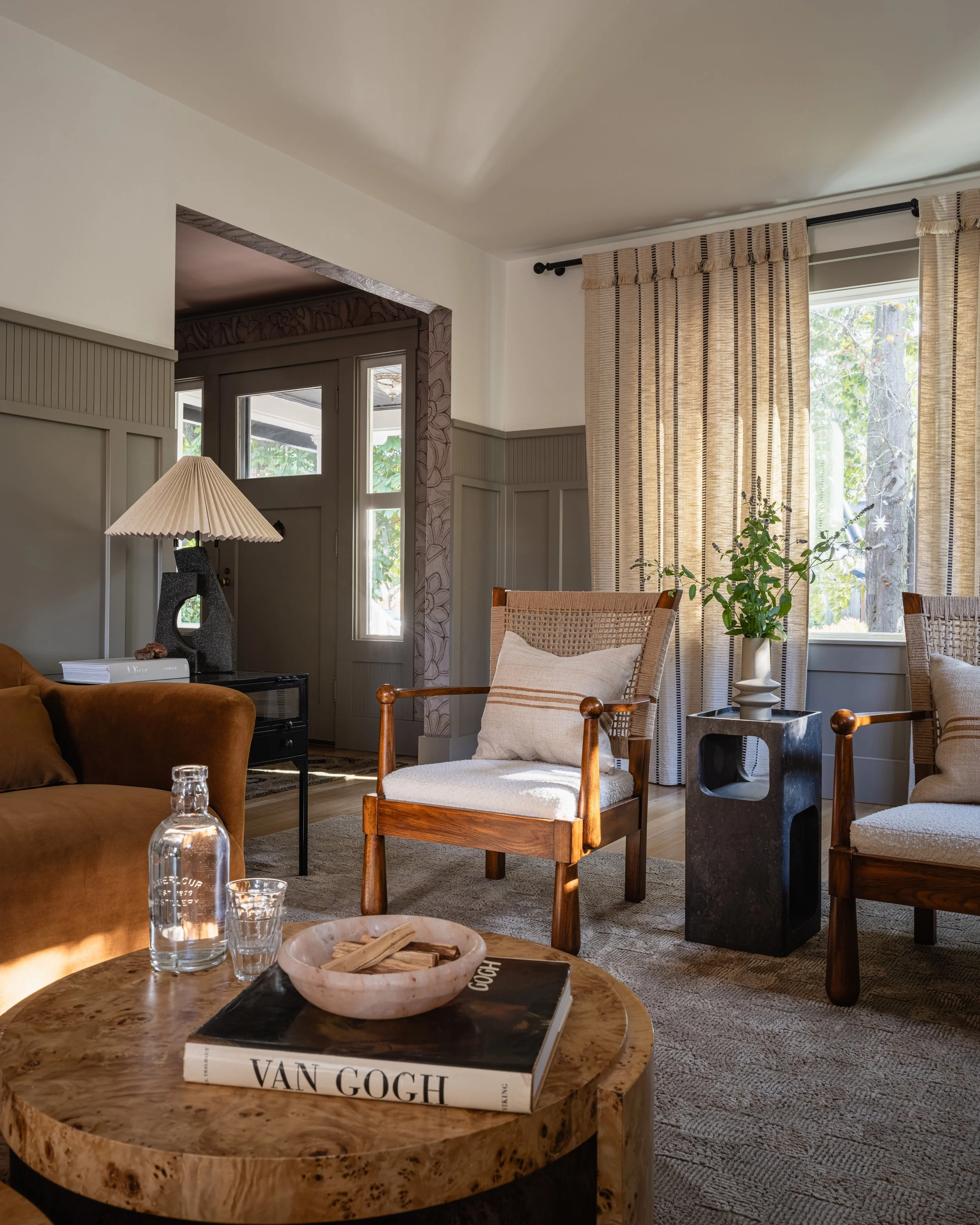Cozy modern living room with wooden chairs and beige cushions, round wooden table with a "Van Gogh" book, a bottle of water, and a decorative dish. A beige sofa, pleated lamp, large windows with curtains, and indoor plants complete the decor.