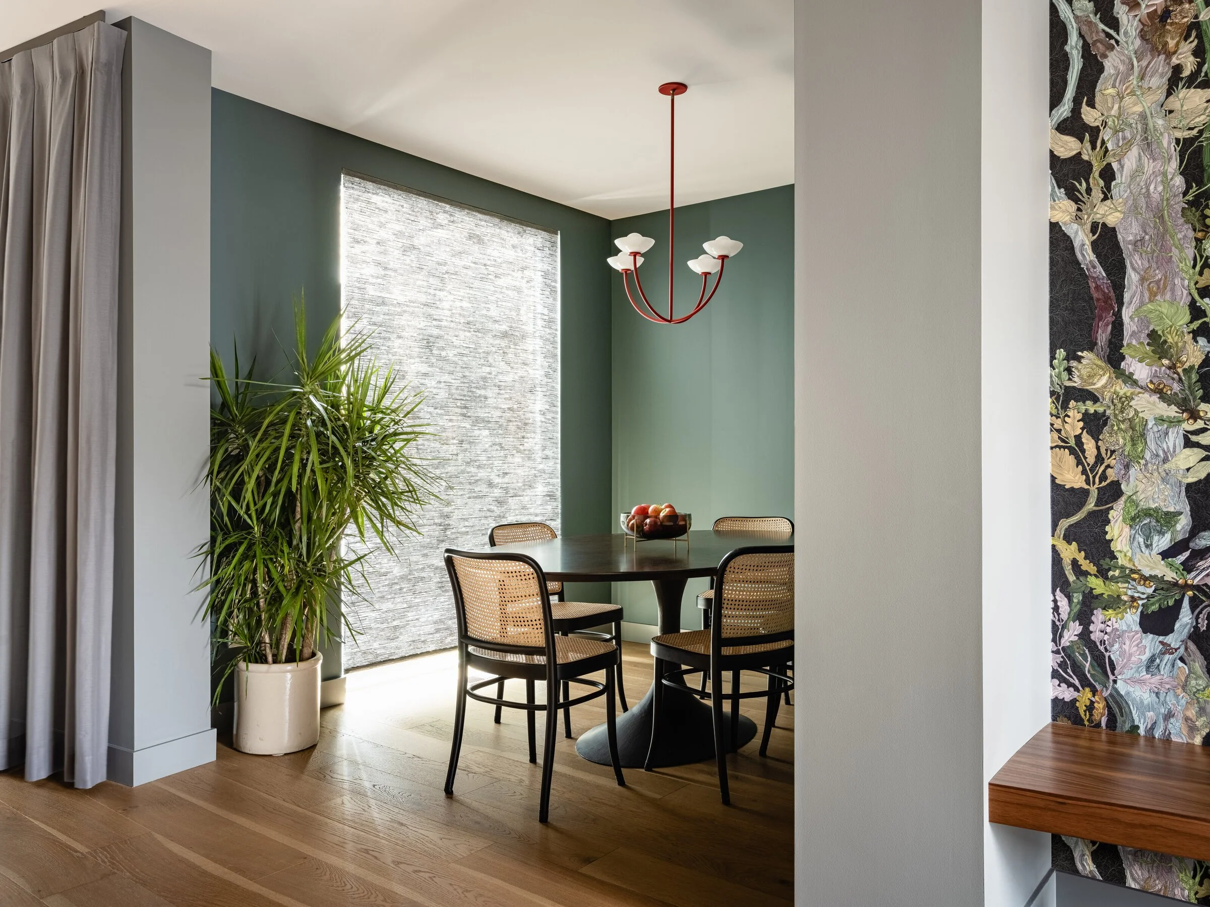 Modern dining room with a round table, four cane-backed chairs, a red chandelier, a potted plant, and decorative wall paneling.