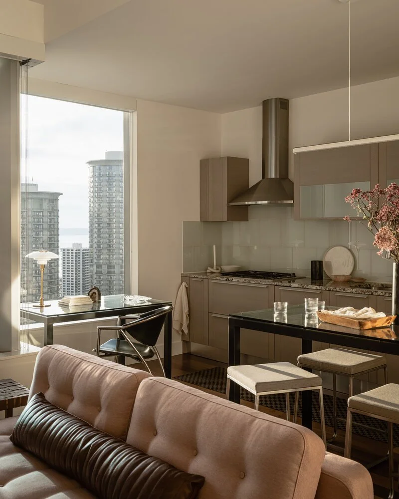 Modern apartment interior with a cozy living room and kitchen, featuring a tufted sofa, glass dining table, bar stools, and urban cityscape view through large windows.