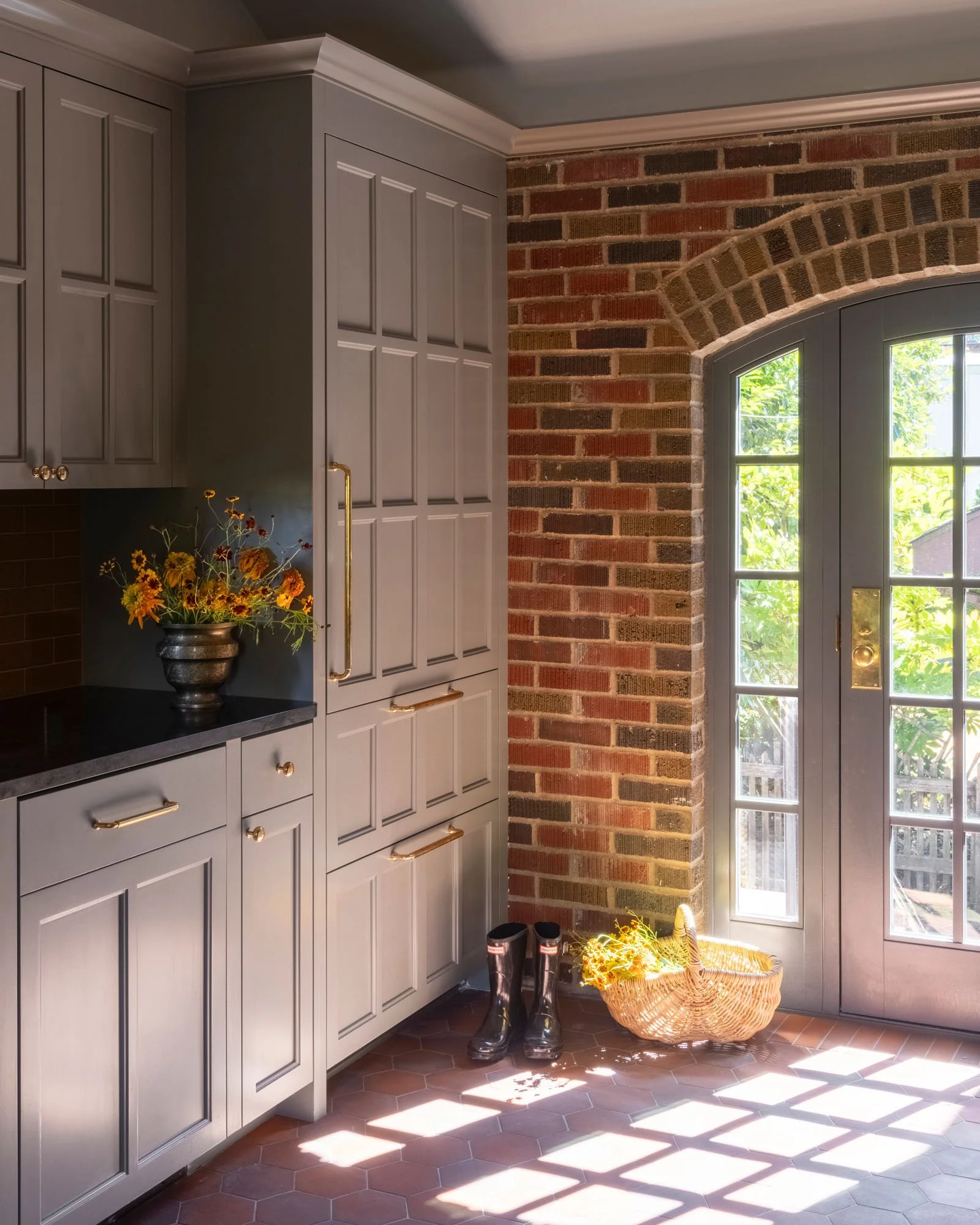 A cozy kitchen corner with gray cabinets, a black countertop, and a vase with yellow and red flowers. There are black rain boots and a woven basket with yellow flowers near a glass-paneled door leading outside, with sunlight creating shadows on the h
