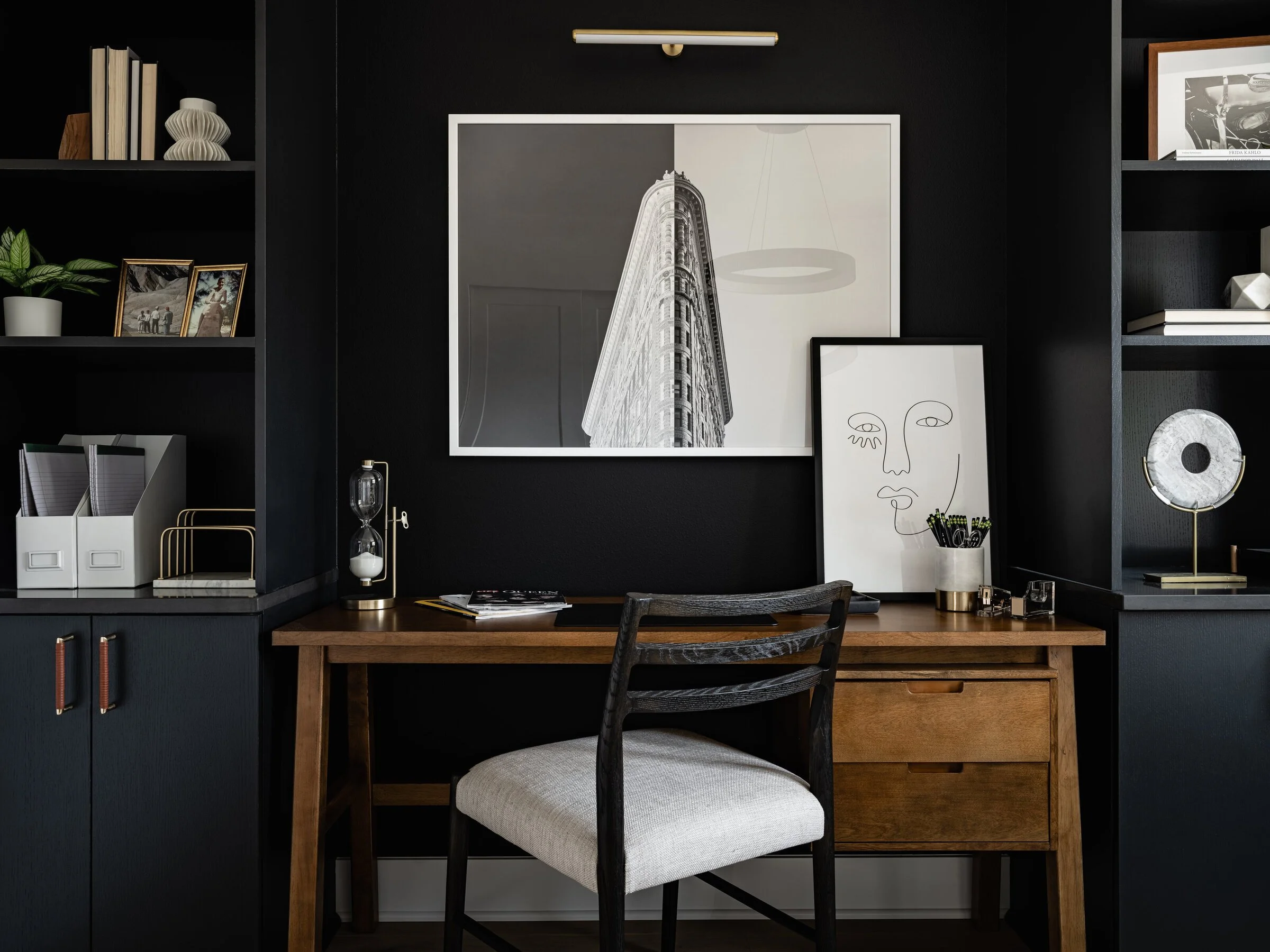 Modern home office with black walls, wooden desk, and abstract wall art. Shelves with books, plants, and decorative items.