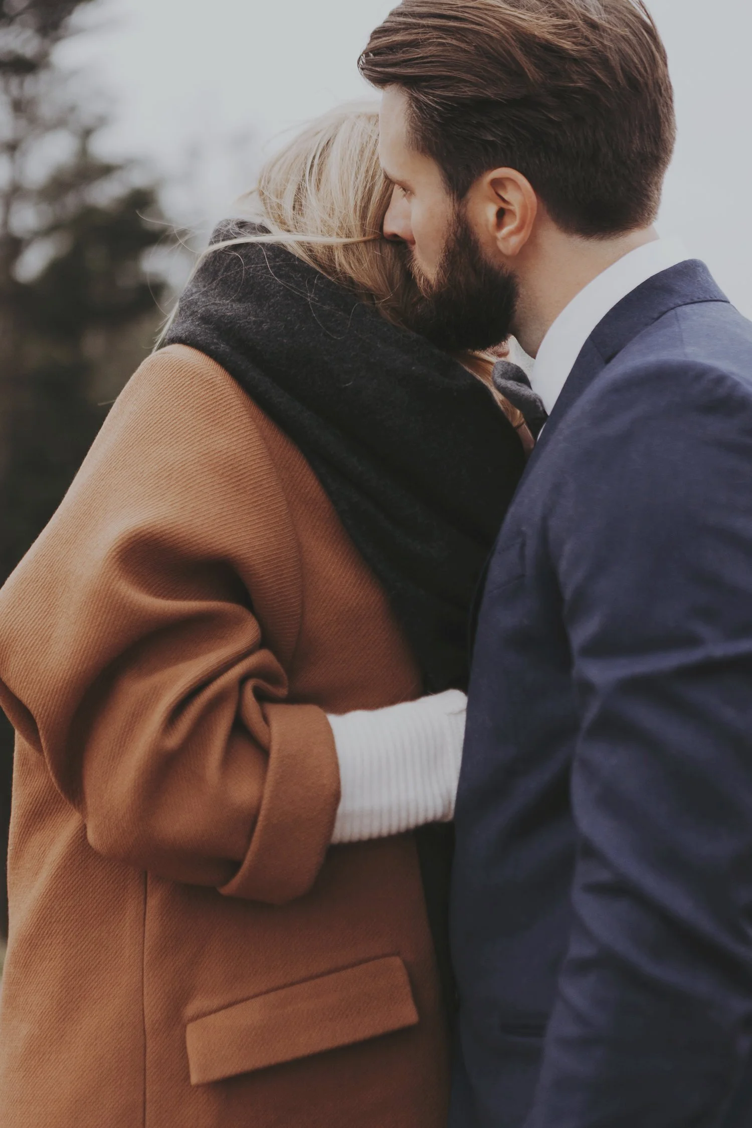 A couple hugging, with the man wearing a blue suit and the woman in a brown coat and dark scarf, outdoors.