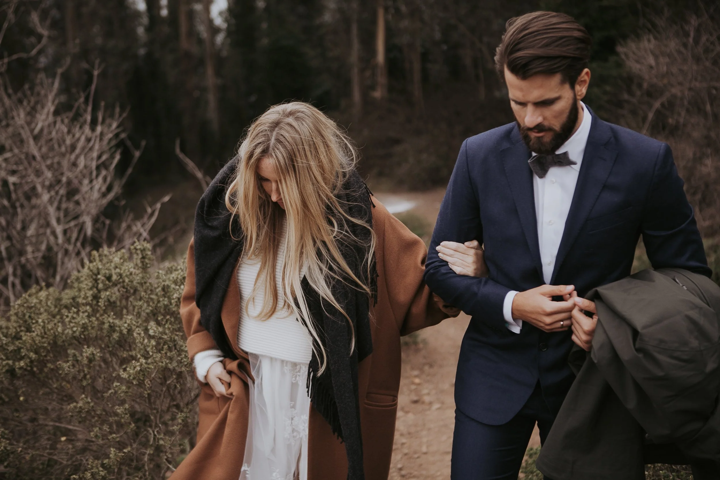 A man in a suit with a bow tie walking arm-in-arm with a woman in a long coat and scarf on a path surrounded by trees.