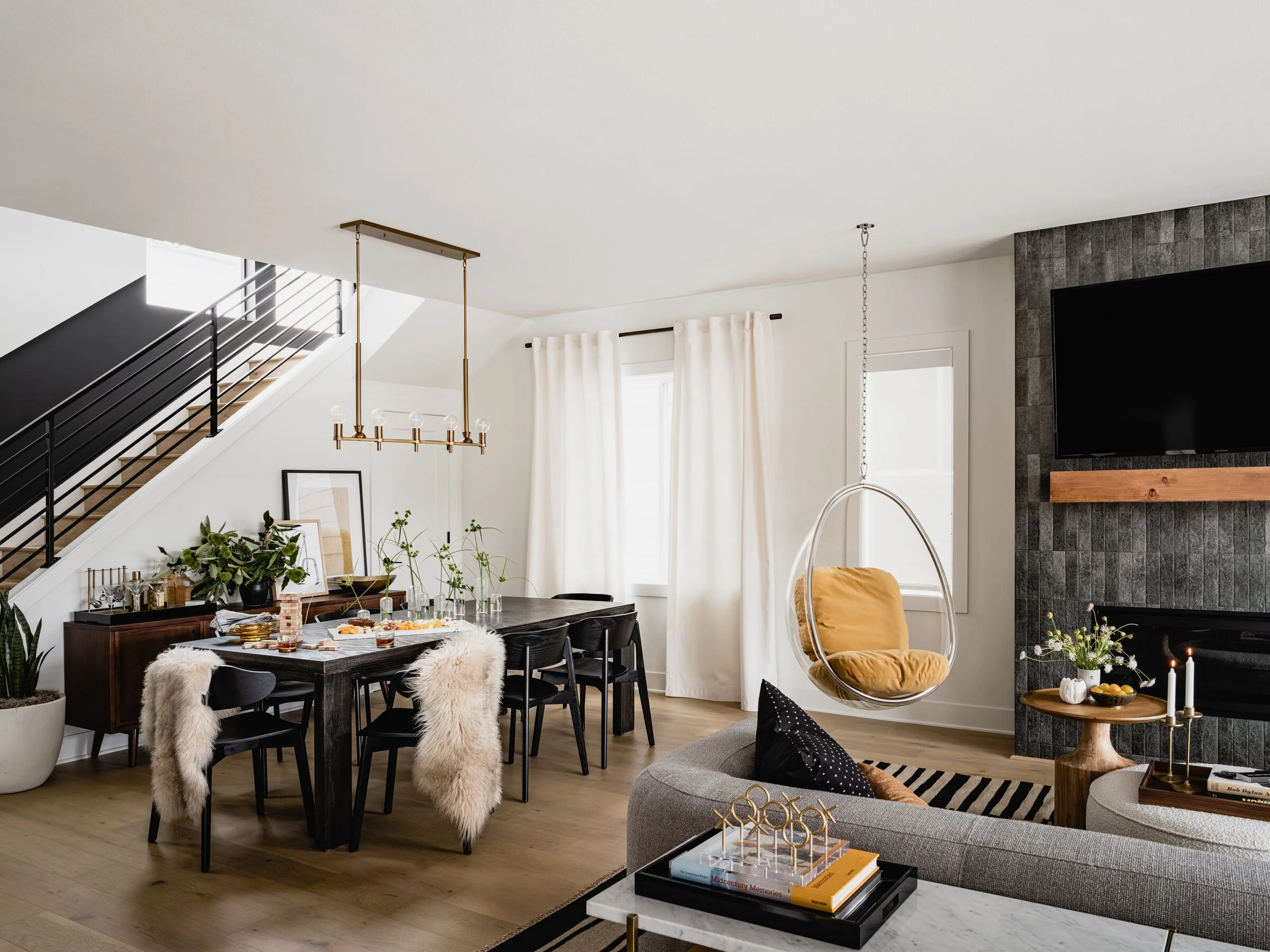 Modern living and dining area with minimalist design, featuring a black dining table with plush chairs draped with faux fur, a cozy gray sofa, a hanging bubble chair, and a wall-mounted TV above a fireplace. Stairs with black railings are visible in