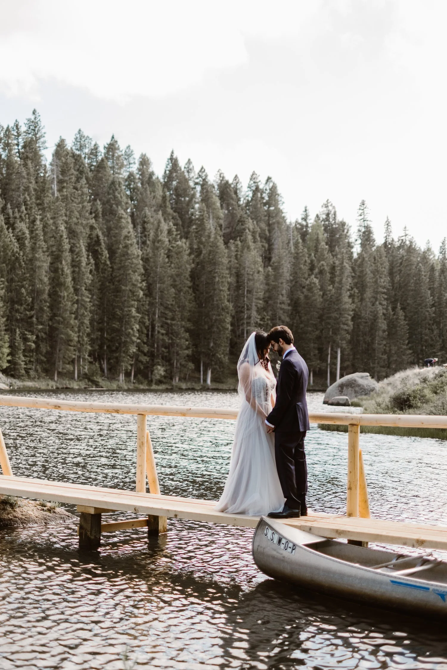 Bride and groom standing on a wooden dock by a lake with a forest background, next to a canoe.