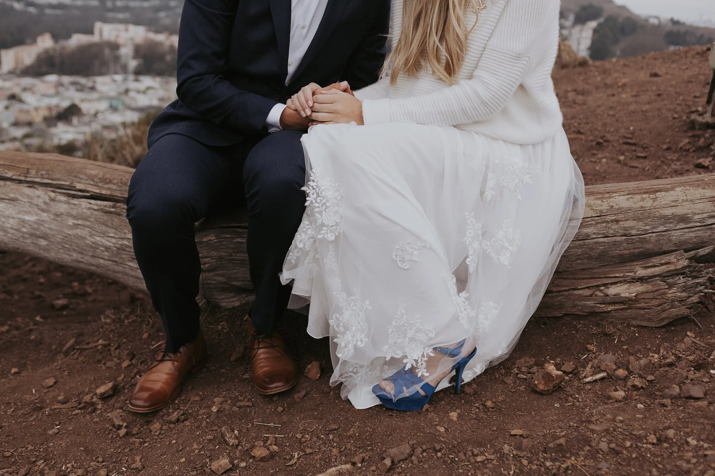 Couple sitting on a log, holding hands; woman in white dress with blue shoes, man in dark suit with brown shoes, outdoor setting.