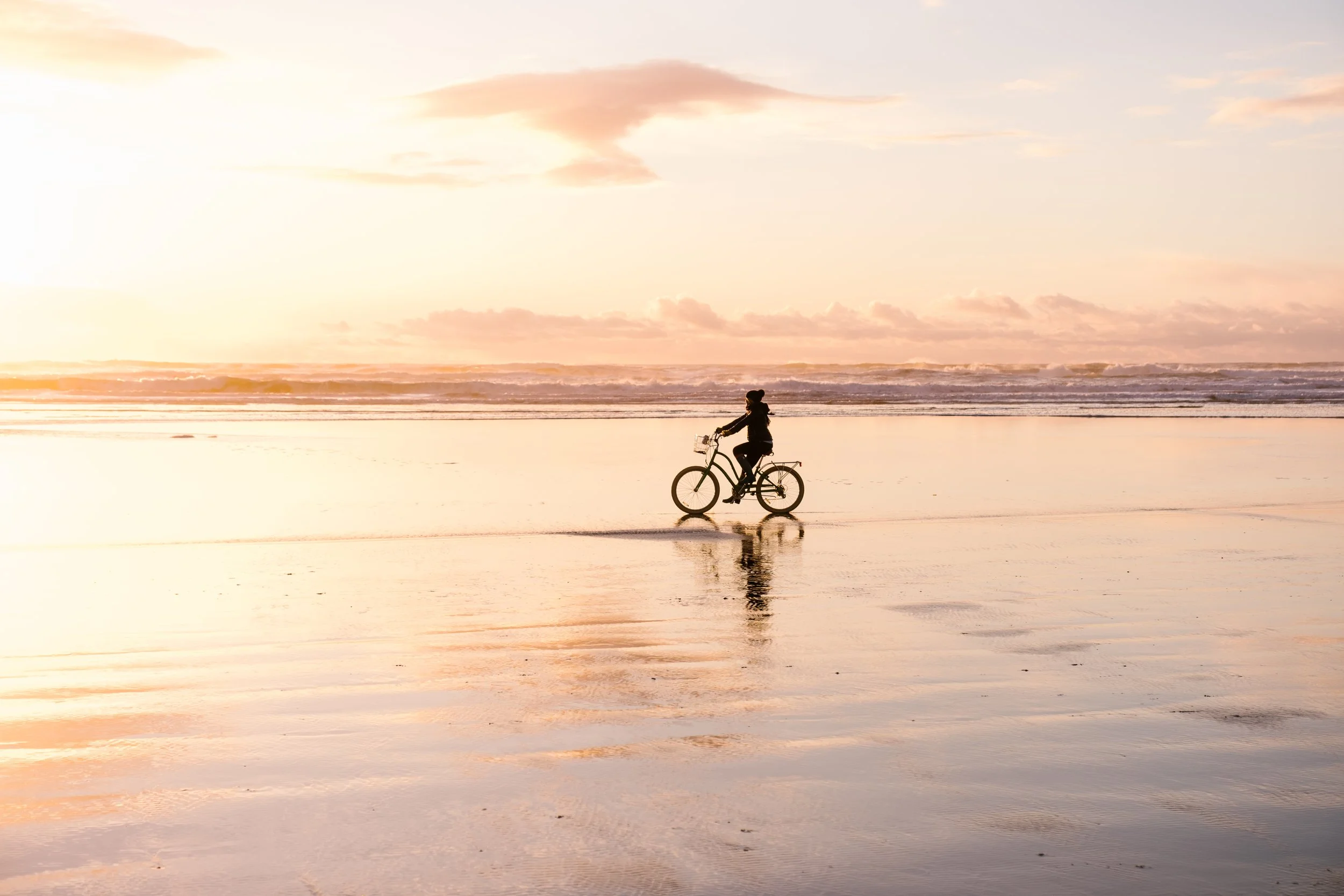 Person riding a bicycle on a beach at sunset with the ocean in the background.